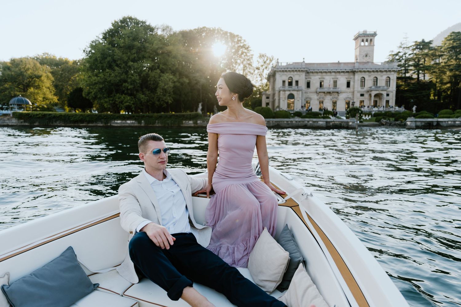 Couple on a wooden boat at golden hour with a historic villa and lush trees reflecting on calm lake waters.