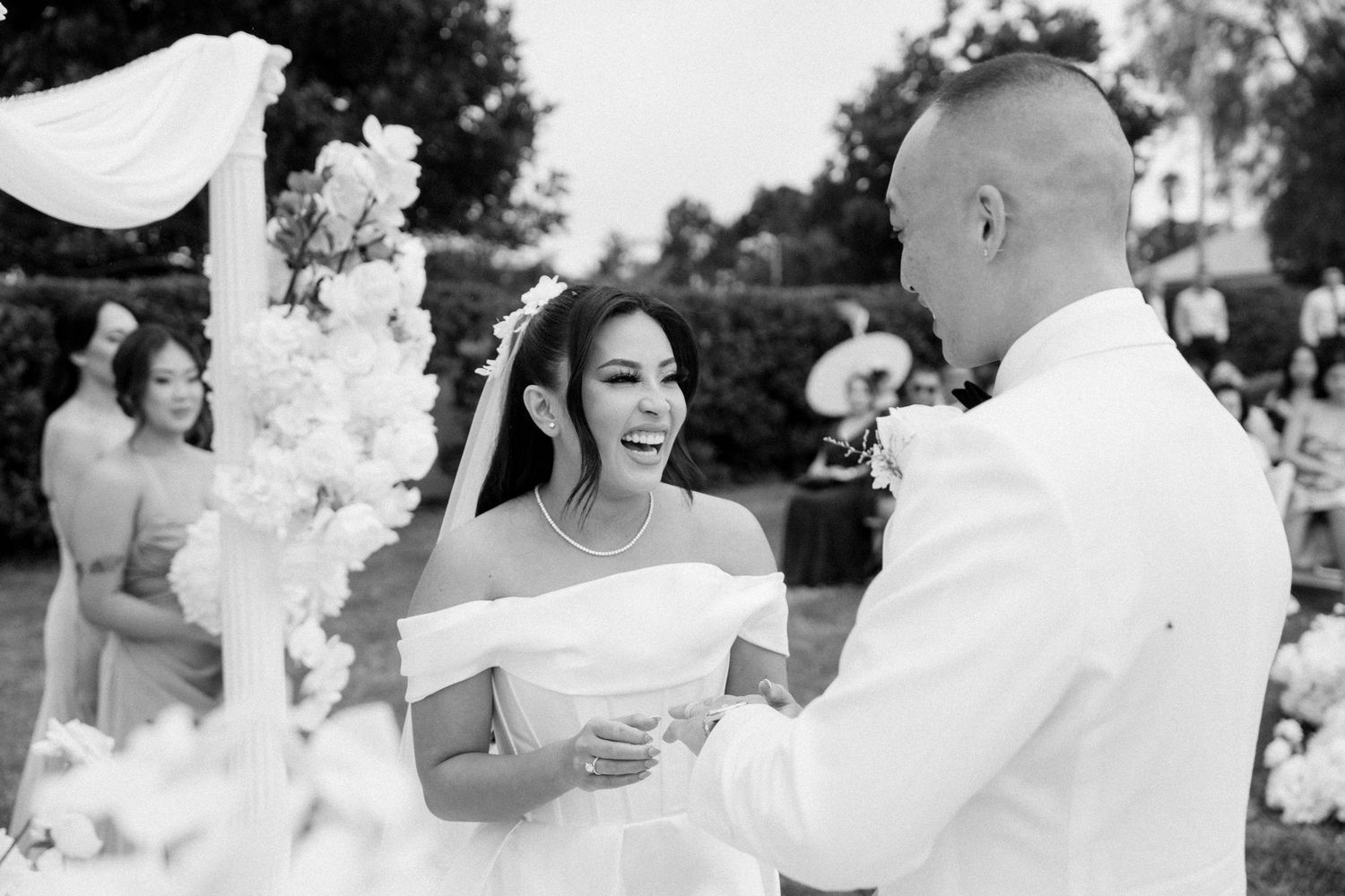 A joyful black and white moment of a bride and groom exchanging vows during an outdoor wedding ceremony.