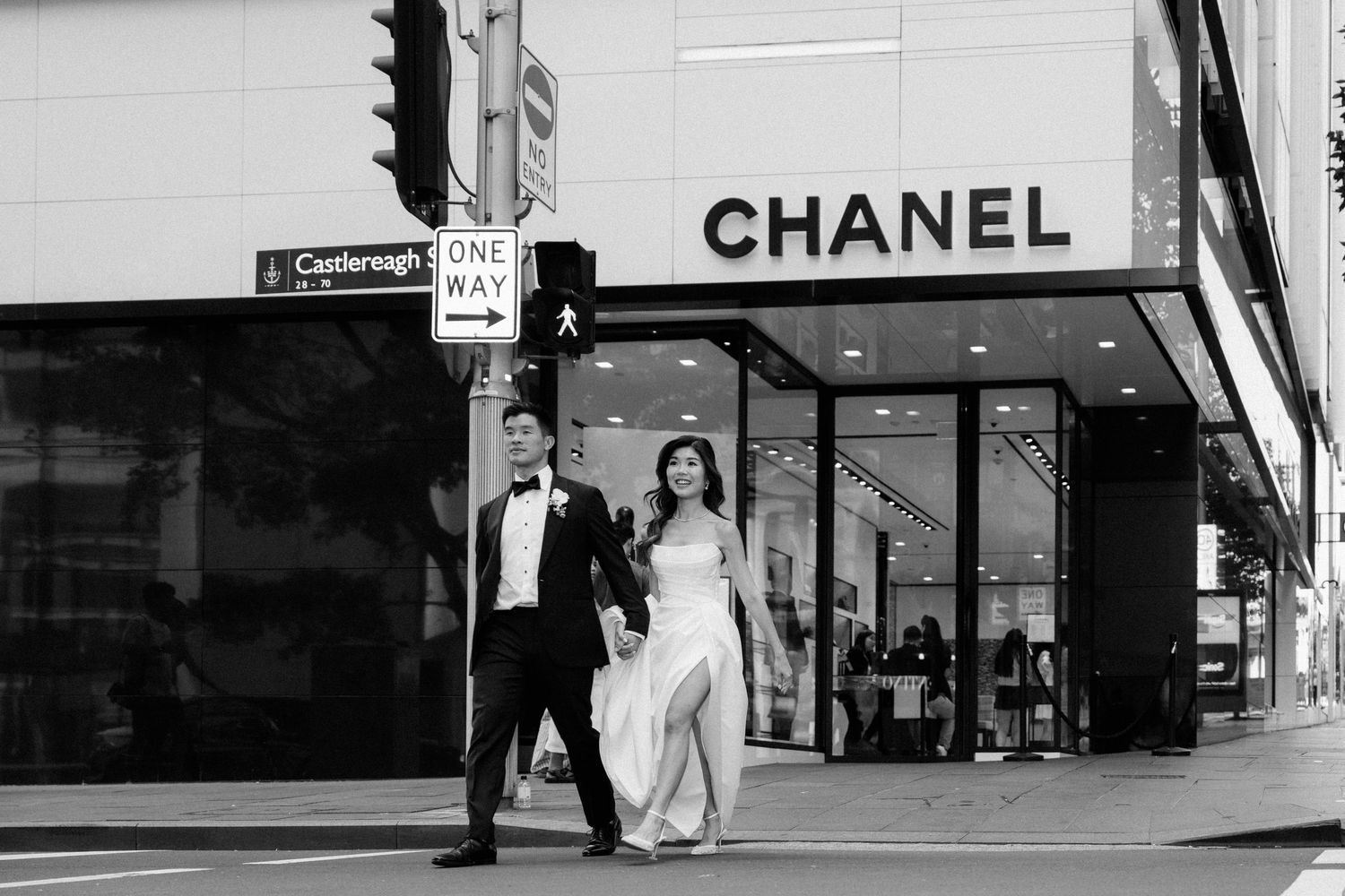 Black and white photo of a couple in formal wedding attire crossing a street in front of a Chanel store.