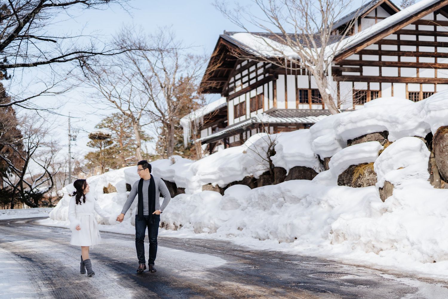 Couple holding hands walking on a snowy road beside a traditional Japanese building in winter.