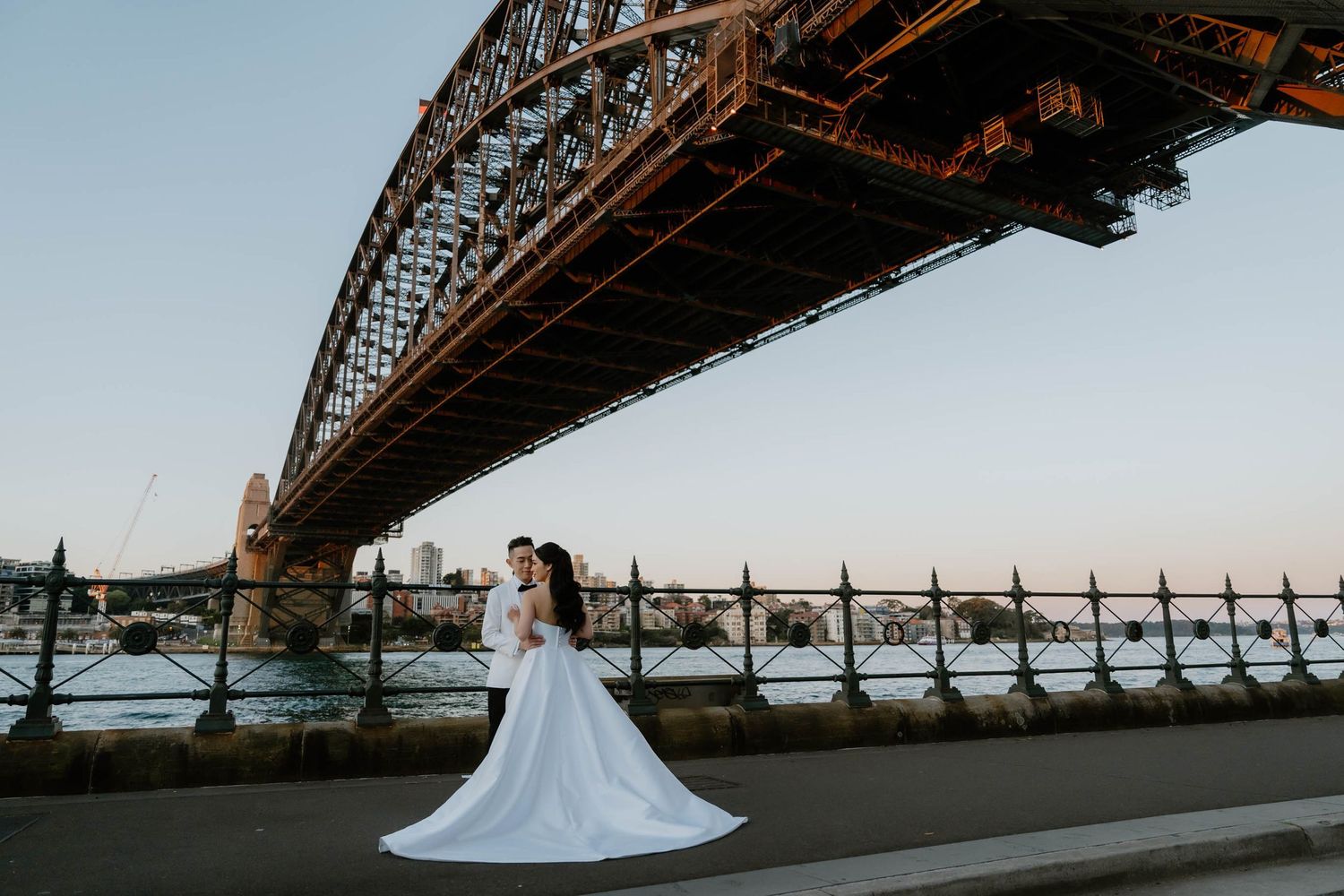 Bride and groom embracing beneath Sydney Harbour Bridge at sunset, with a flowing white wedding gown trailing on the pavement.