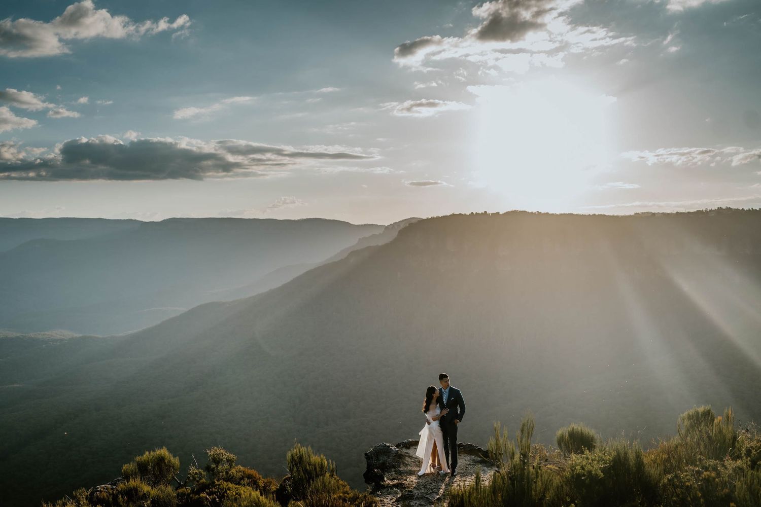 Couple embracing on mountain peak at sunset with dramatic rays of light illuminating vast valley below.