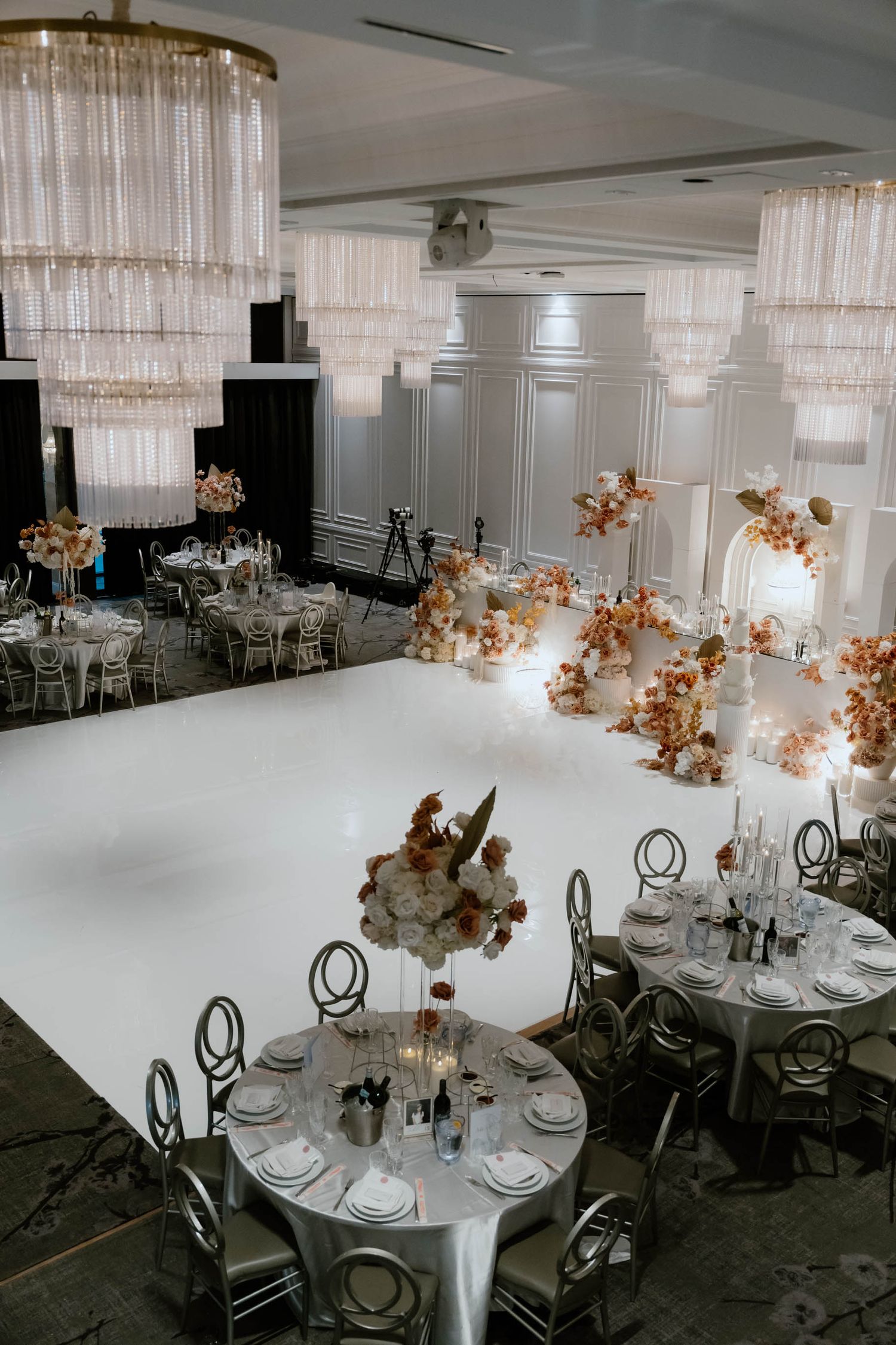 Elegant wedding reception venue with crystal chandeliers, white tables, and peach floral arrangements in a modern ballroom setting.