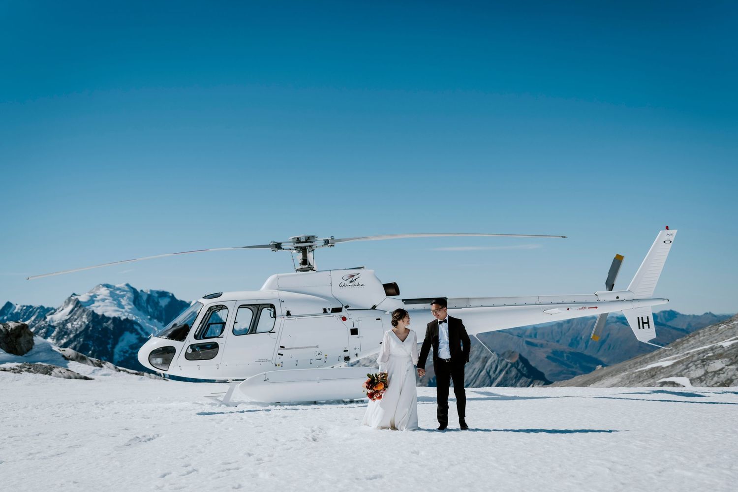 Bride and groom pose on snowy mountain peak beside white helicopter with scenic alpine backdrop.