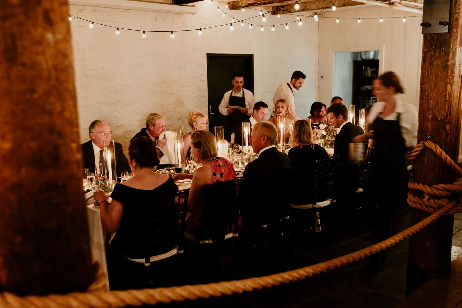 A warmly lit rustic event space with exposed wooden beams and string lights creating an intimate atmosphere for guests.