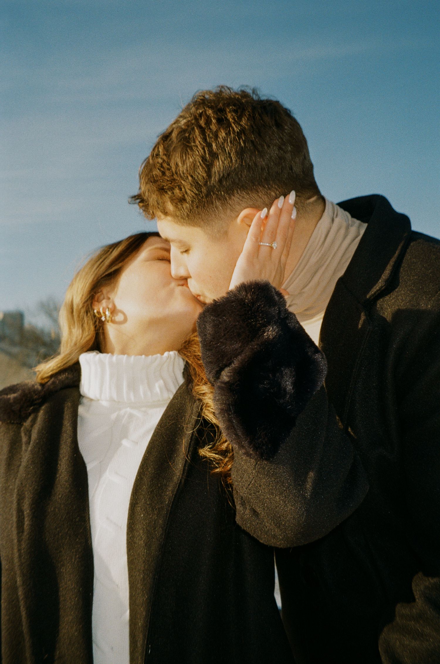 A couple shares a romantic kiss outdoors on a sunny day, dressed in cozy winter coats with a blue sky backdrop.