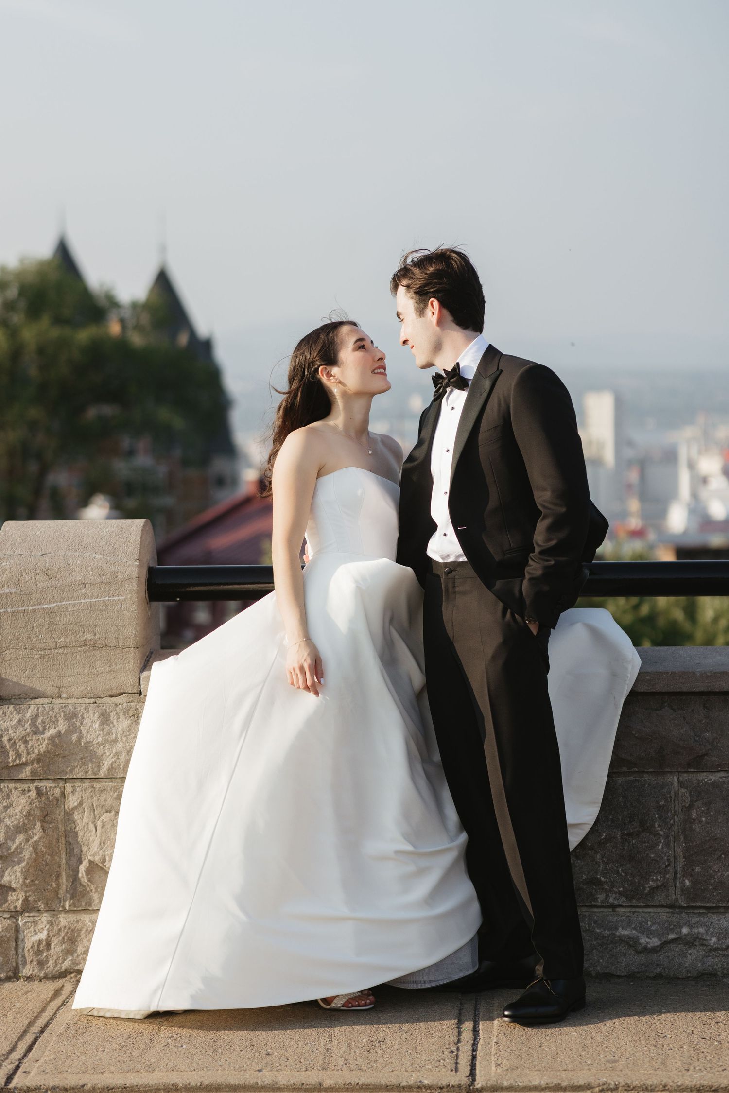 Bride in white strapless gown and groom in black tuxedo share a romantic moment on a terrace with city views.
