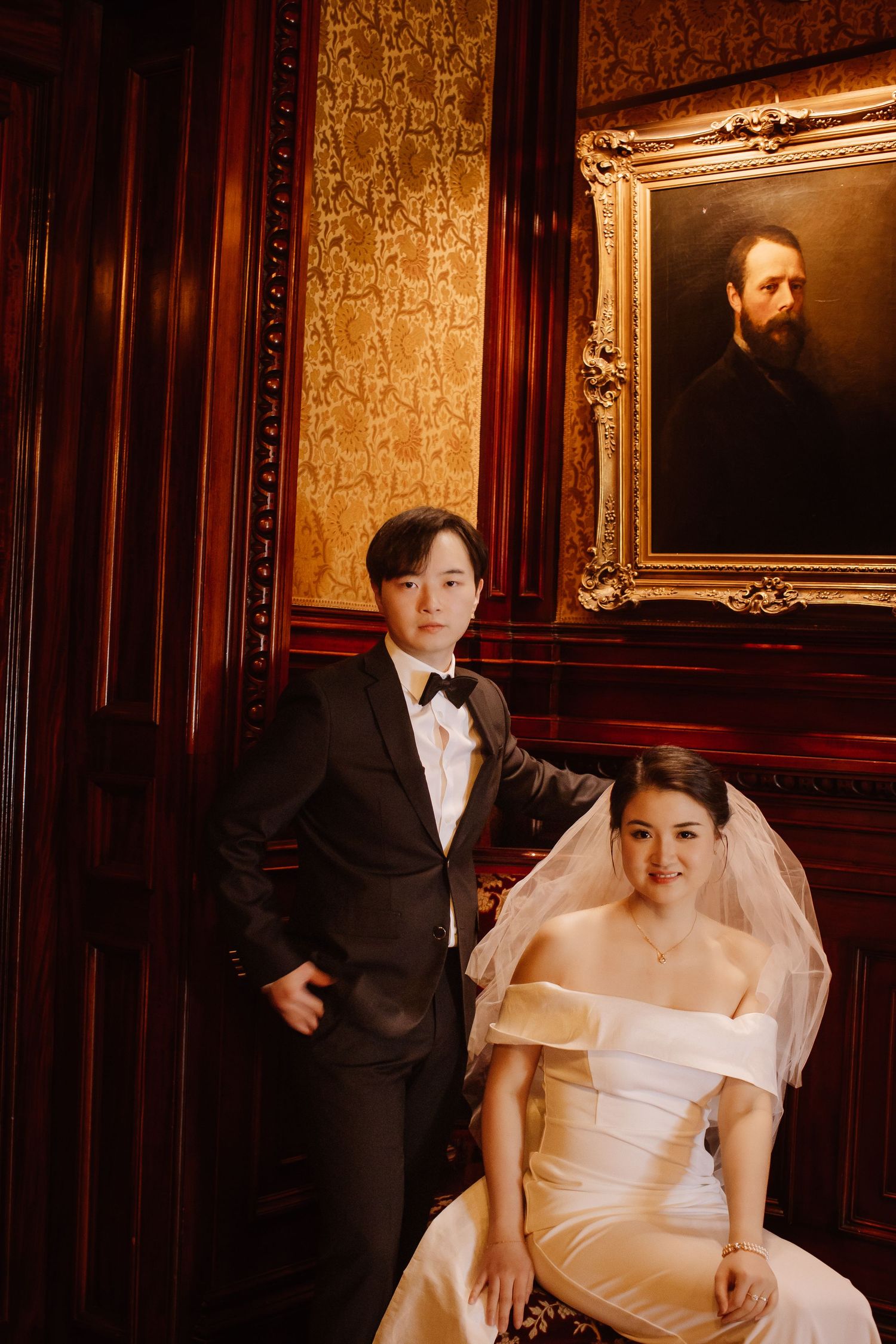 Elegant wedding couple poses in formal attire against dark wood paneled wall with ornate gold-framed portrait.