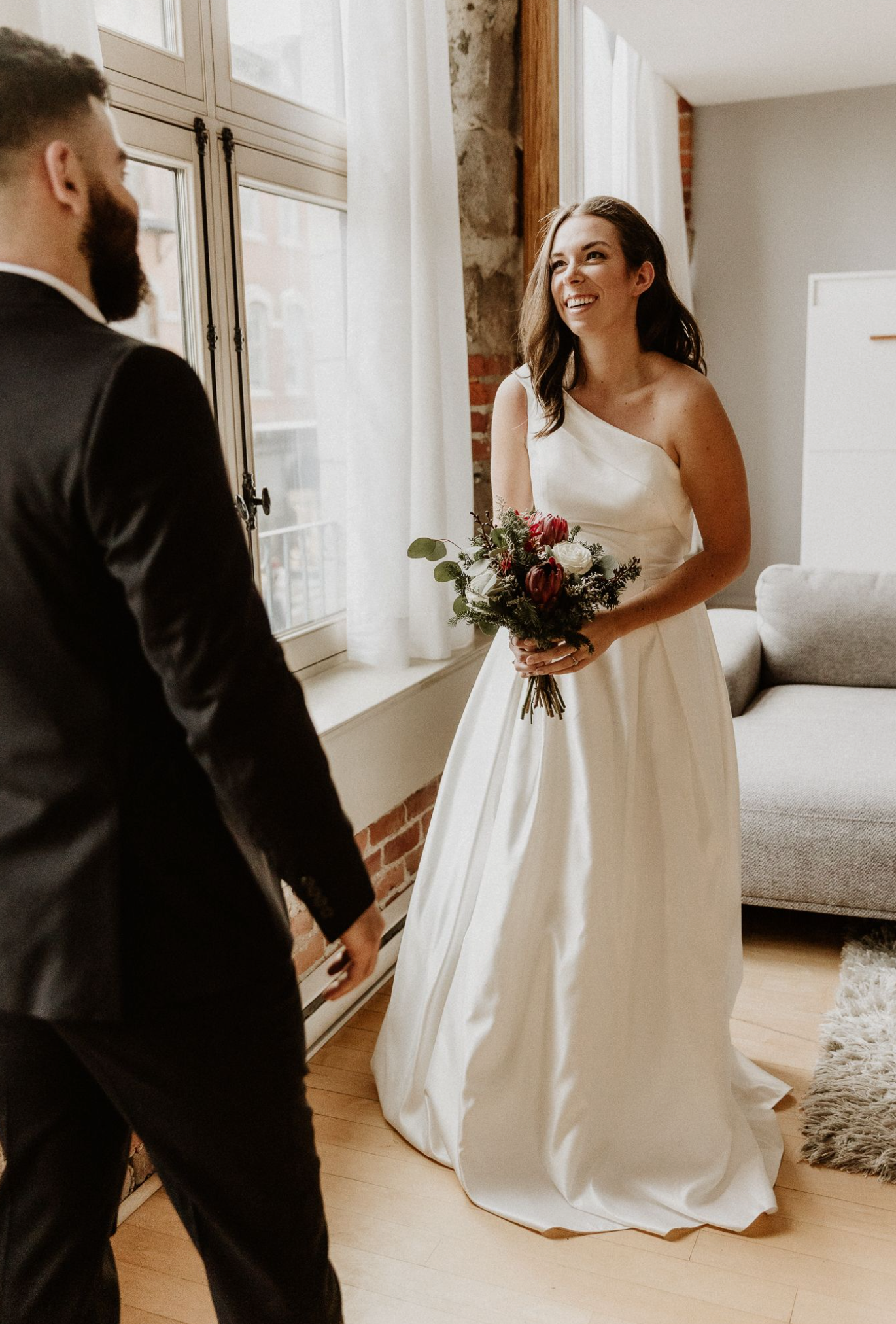 Bride in white one-shoulder gown holding bouquet smiles at groom in black tuxedo during wedding first look.