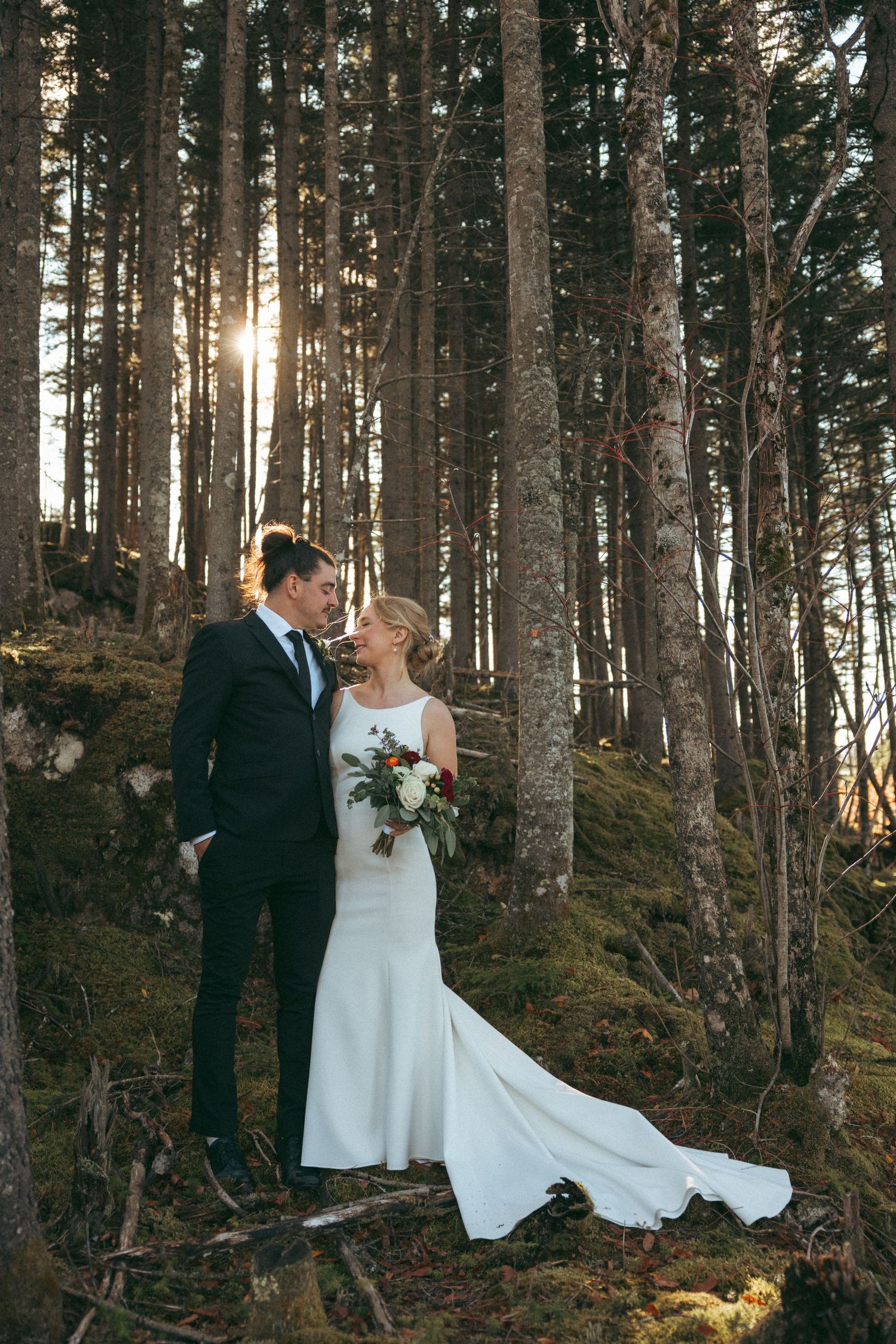 Bride and groom share a romantic moment in a sunlit forest, she holds a bouquet in her elegant white gown.