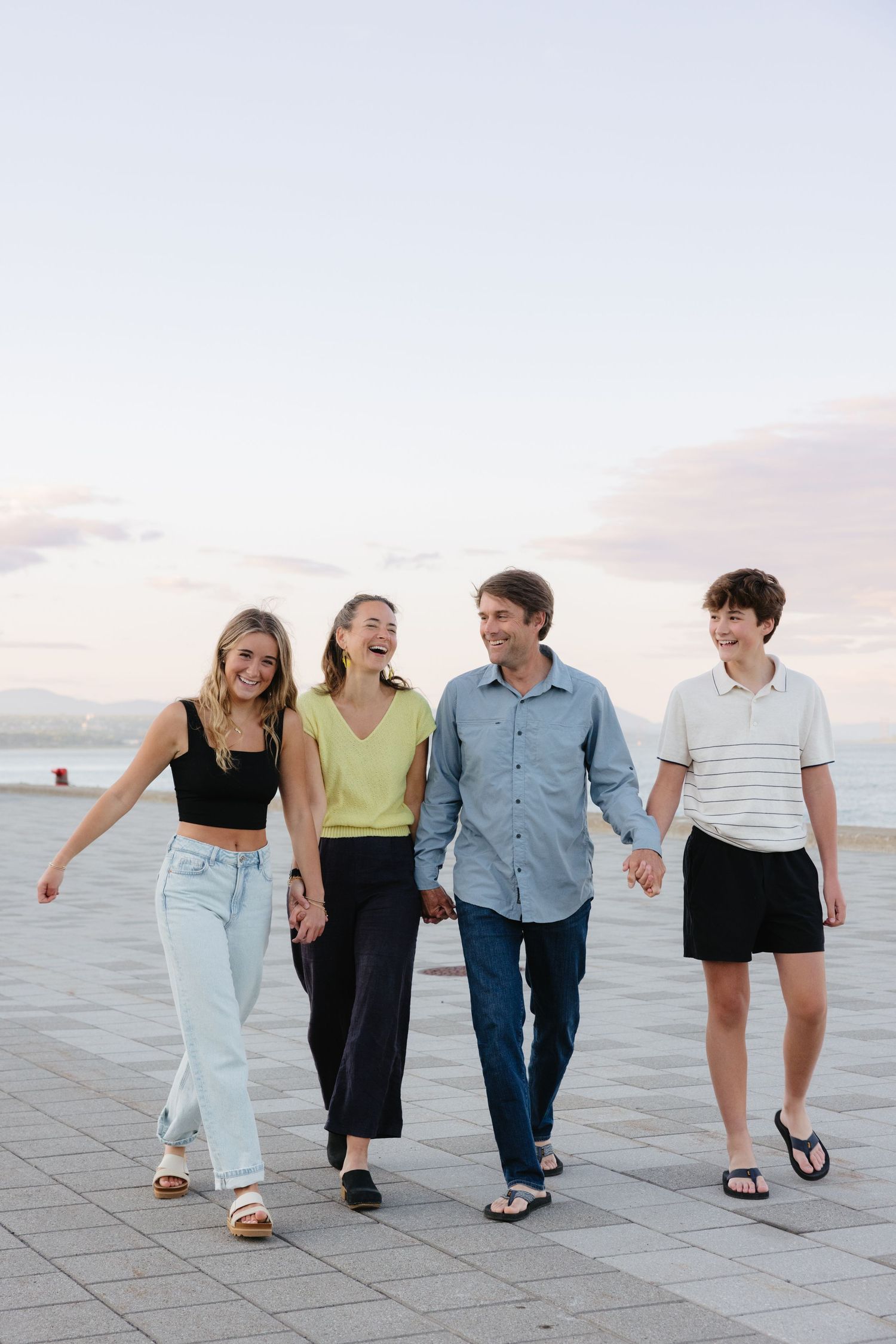 Four young adults walking together on a waterfront promenade at sunset, dressed casually in summer clothing.