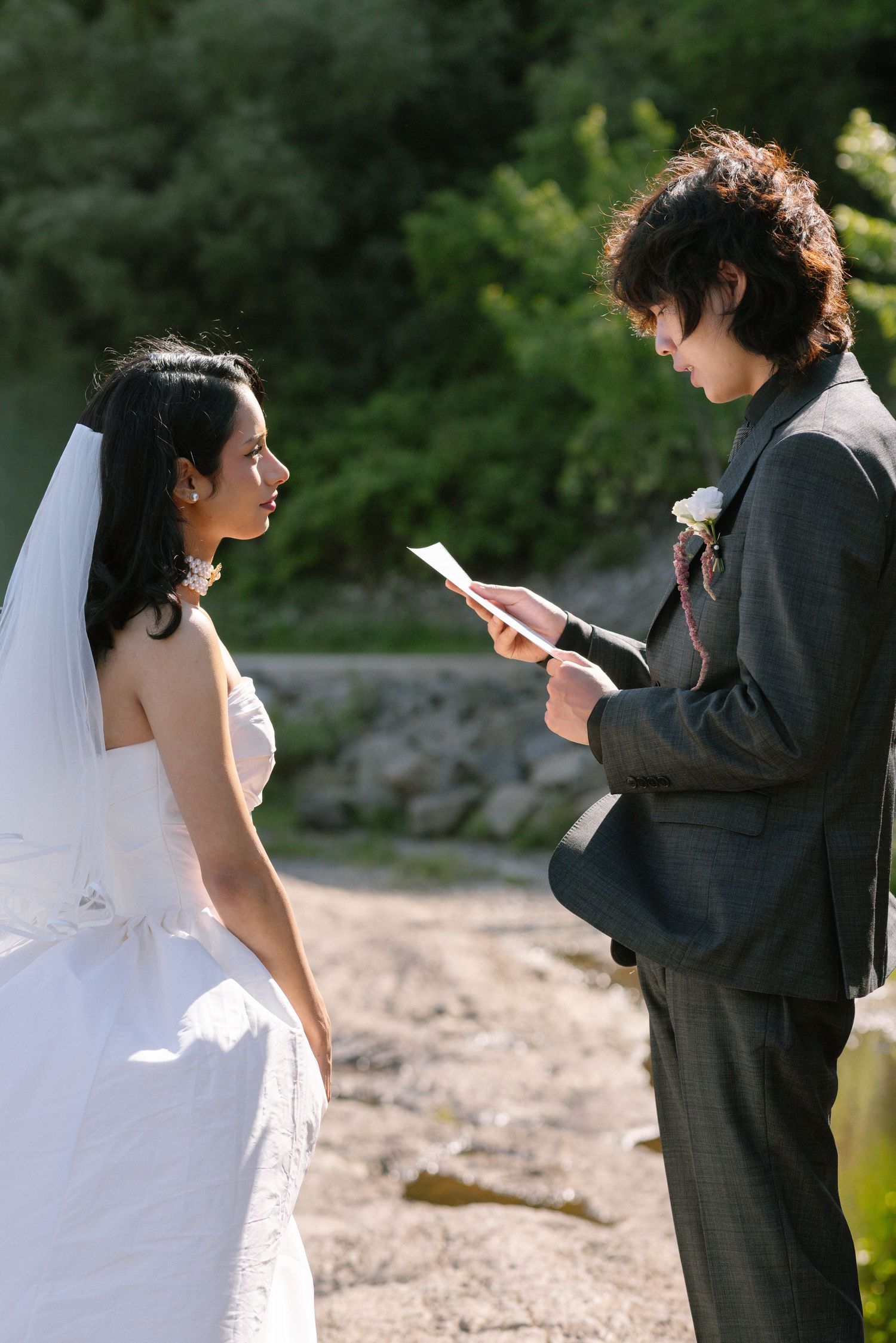 Groom reads wedding vows from paper to bride in white dress and veil during outdoor ceremony.