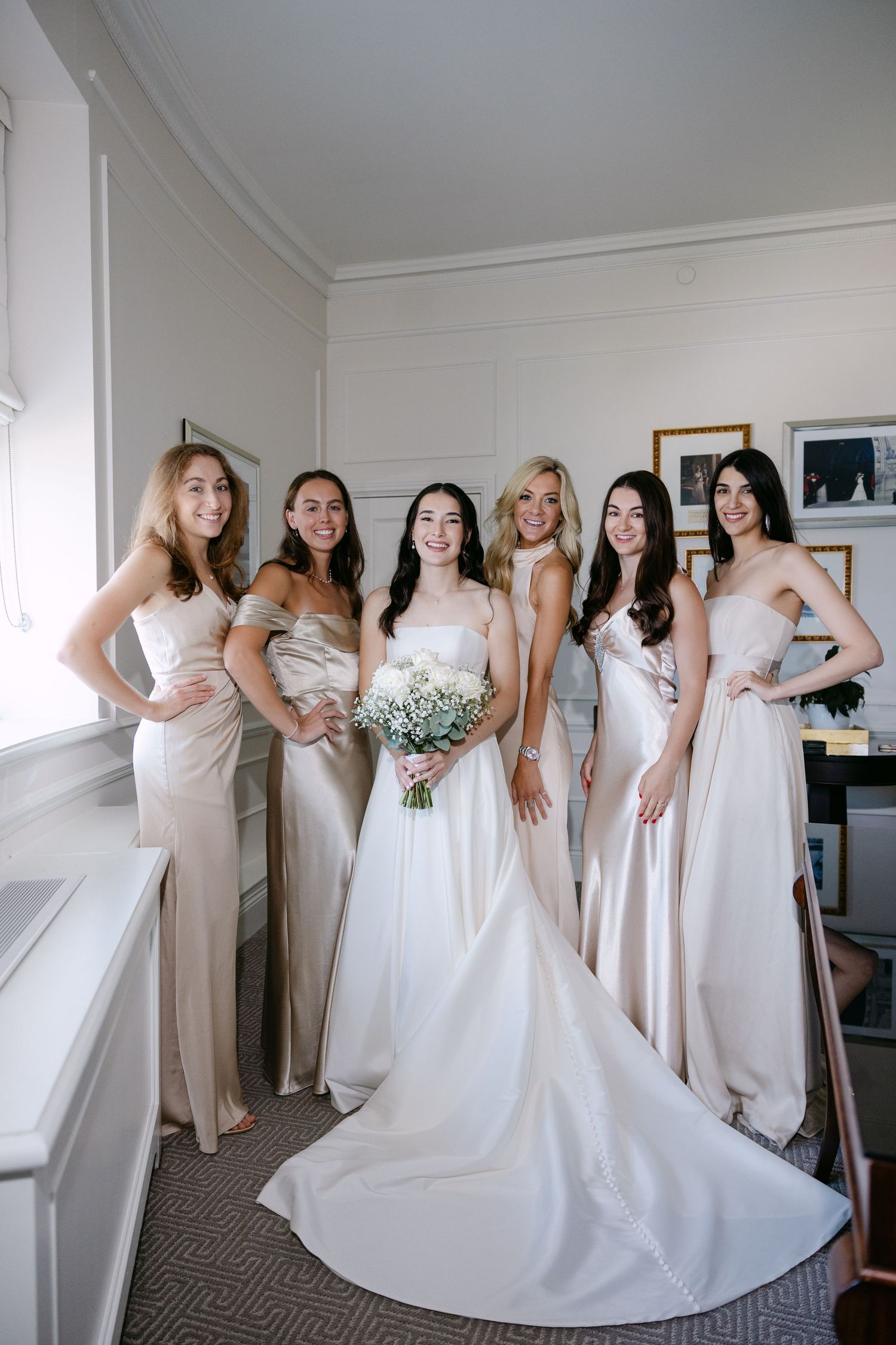 Bride in white gown with five bridesmaids in champagne dresses holding bouquet, posing together in bright room.