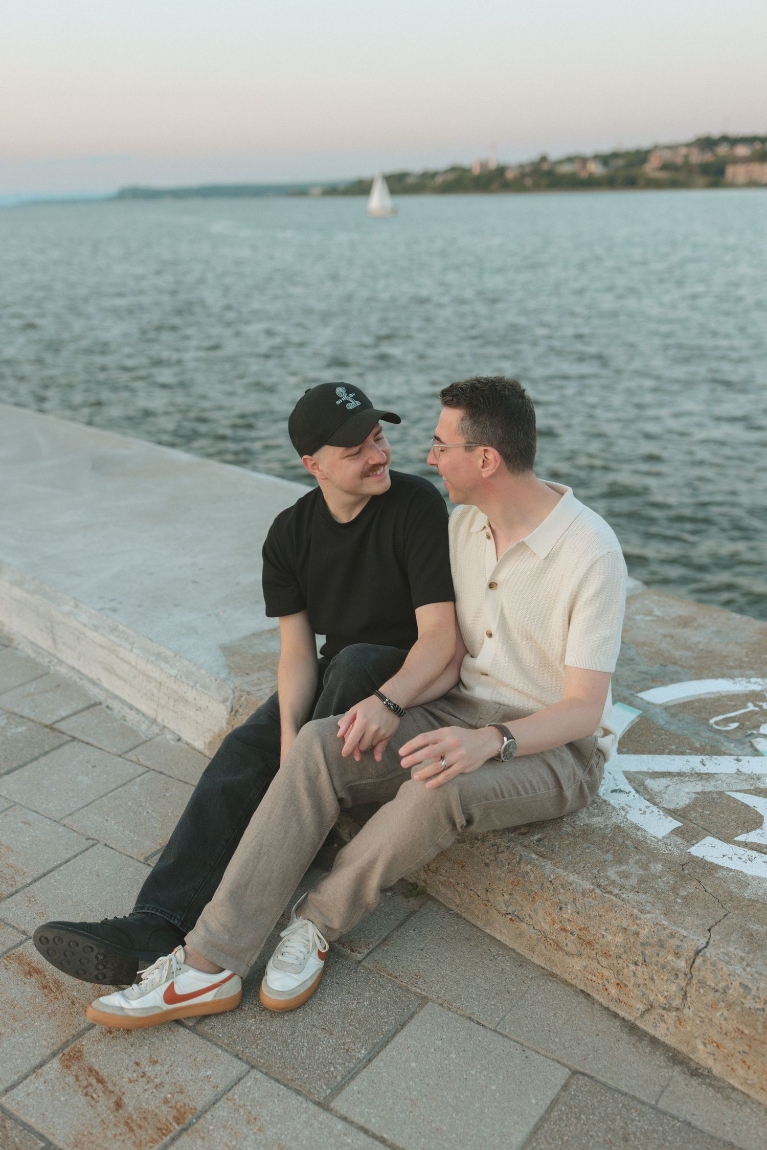 Two men sitting together on a waterfront promenade at sunset, sharing a romantic moment by the sea.