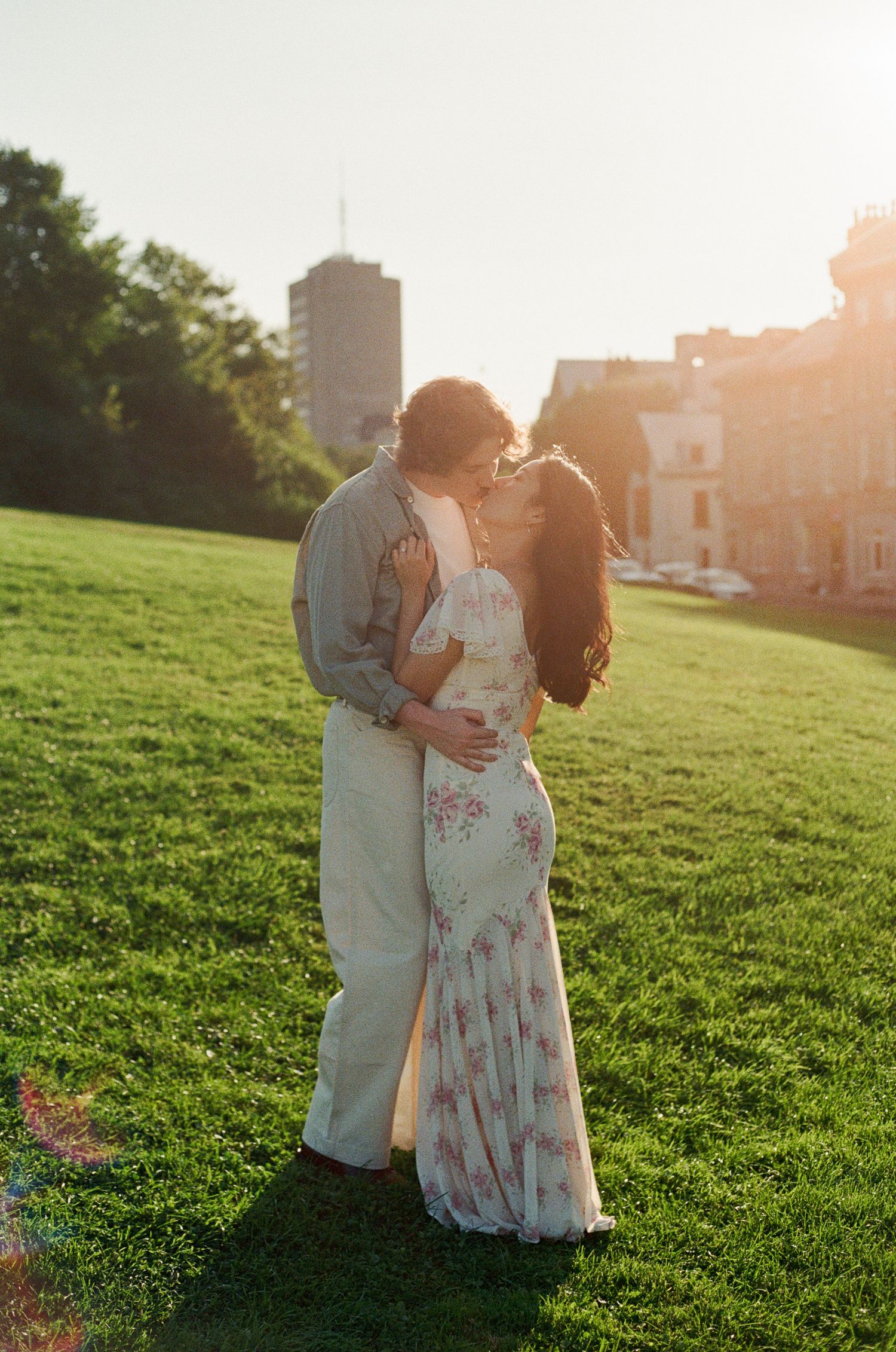 Couple sharing a romantic kiss at golden hour on a lush green lawn with city buildings in the background.