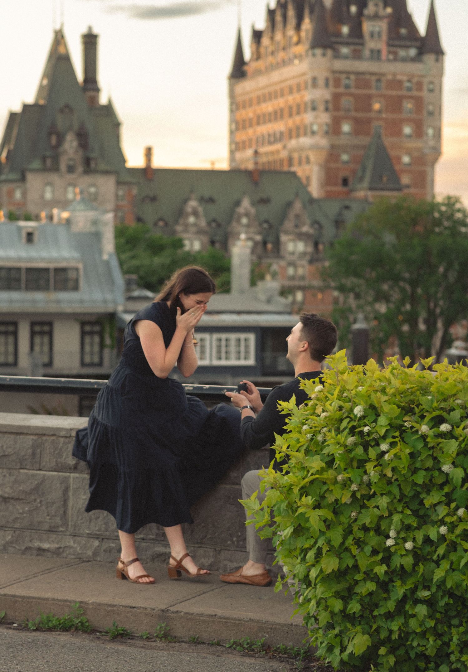 Man proposes to woman on one knee in Quebec City with Chateau Frontenac in the background at sunset.