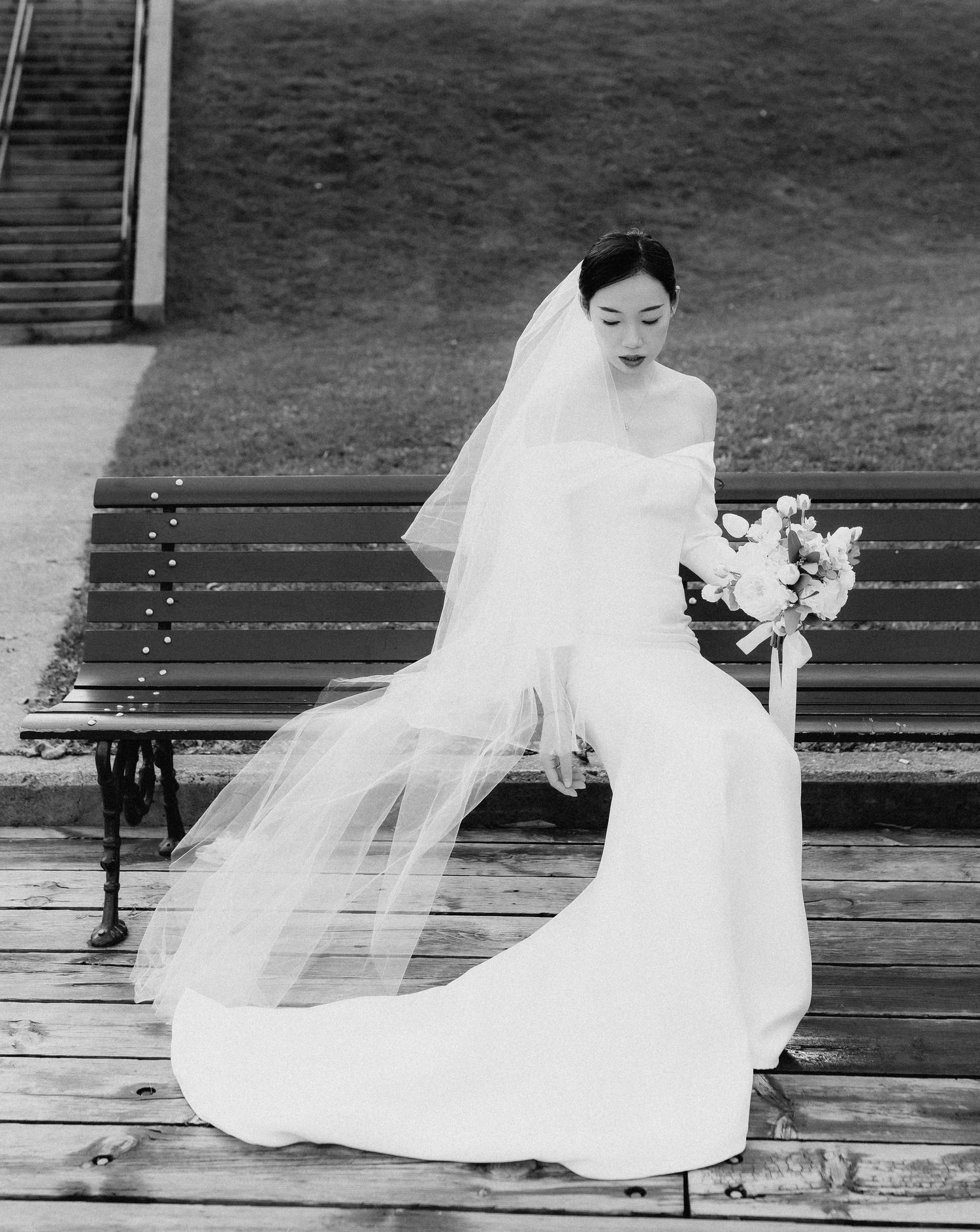 Bride in elegant off-shoulder gown and veil sits on park bench holding bouquet, black and white wedding portrait.