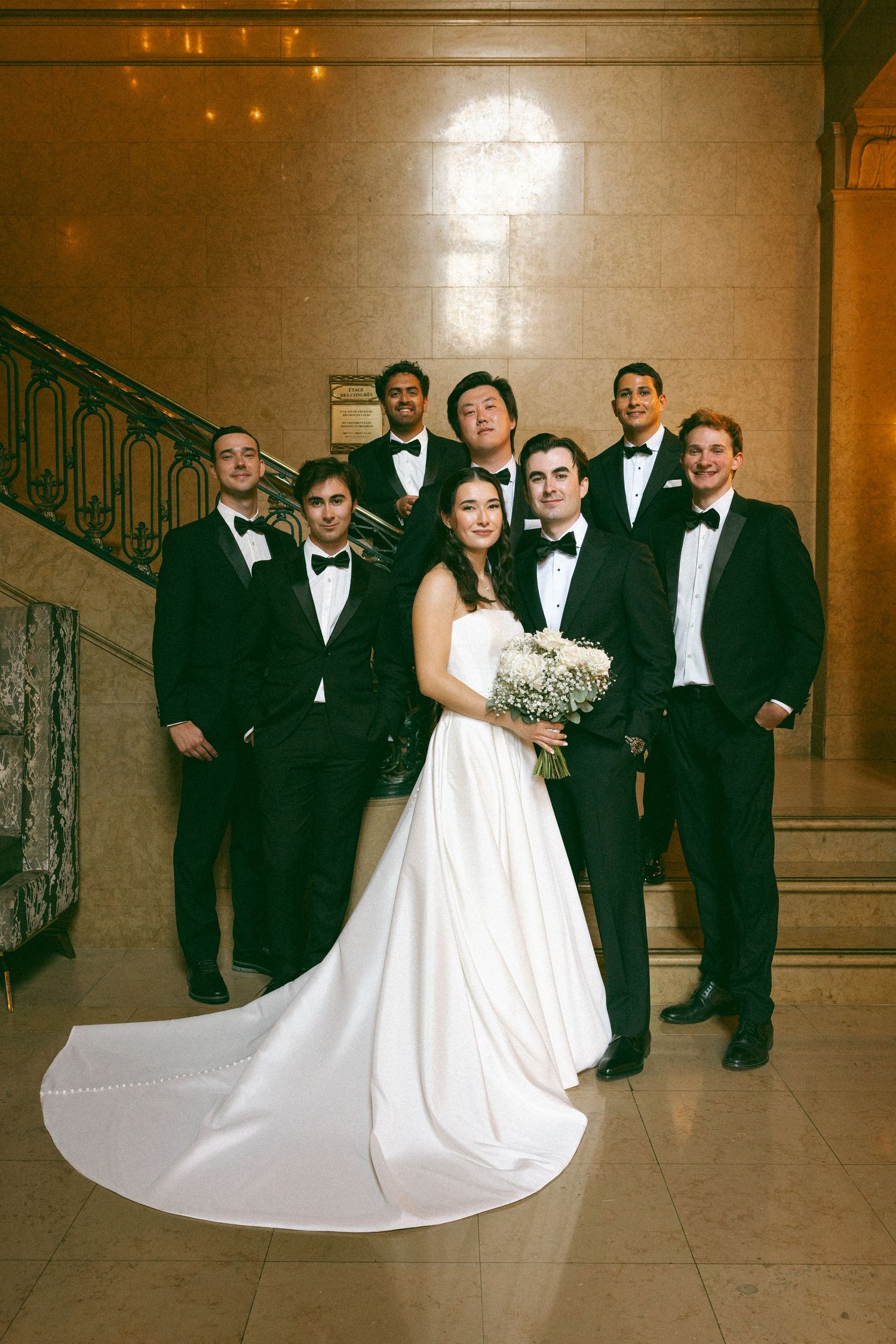 Bride in white gown holds bouquet with groom and groomsmen in black tuxedos posing on marble staircase.