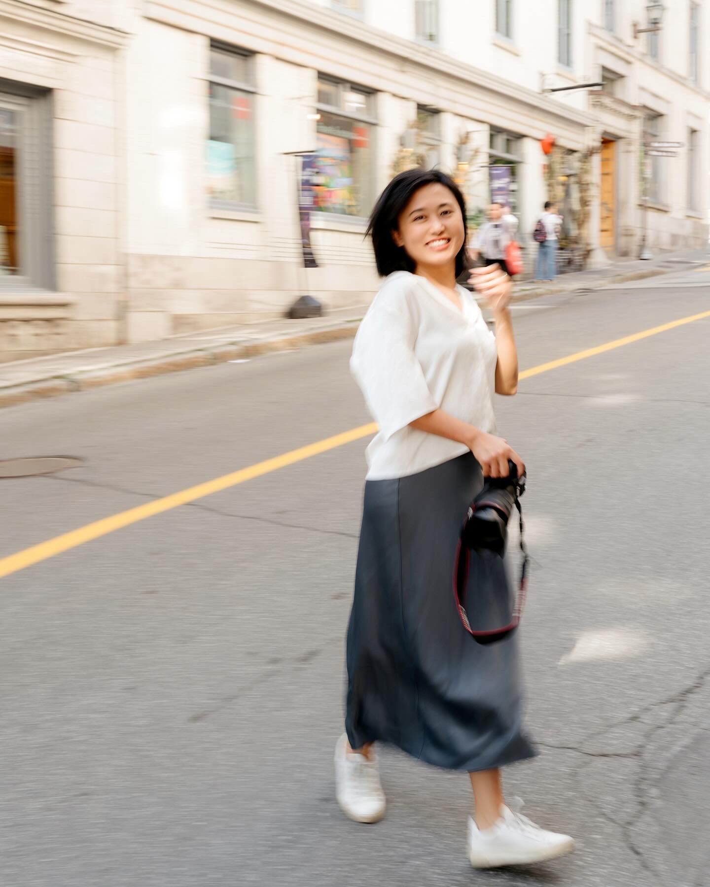 Smiling young Asian woman in white top and gray midi skirt walking on a city street carrying a camera bag.