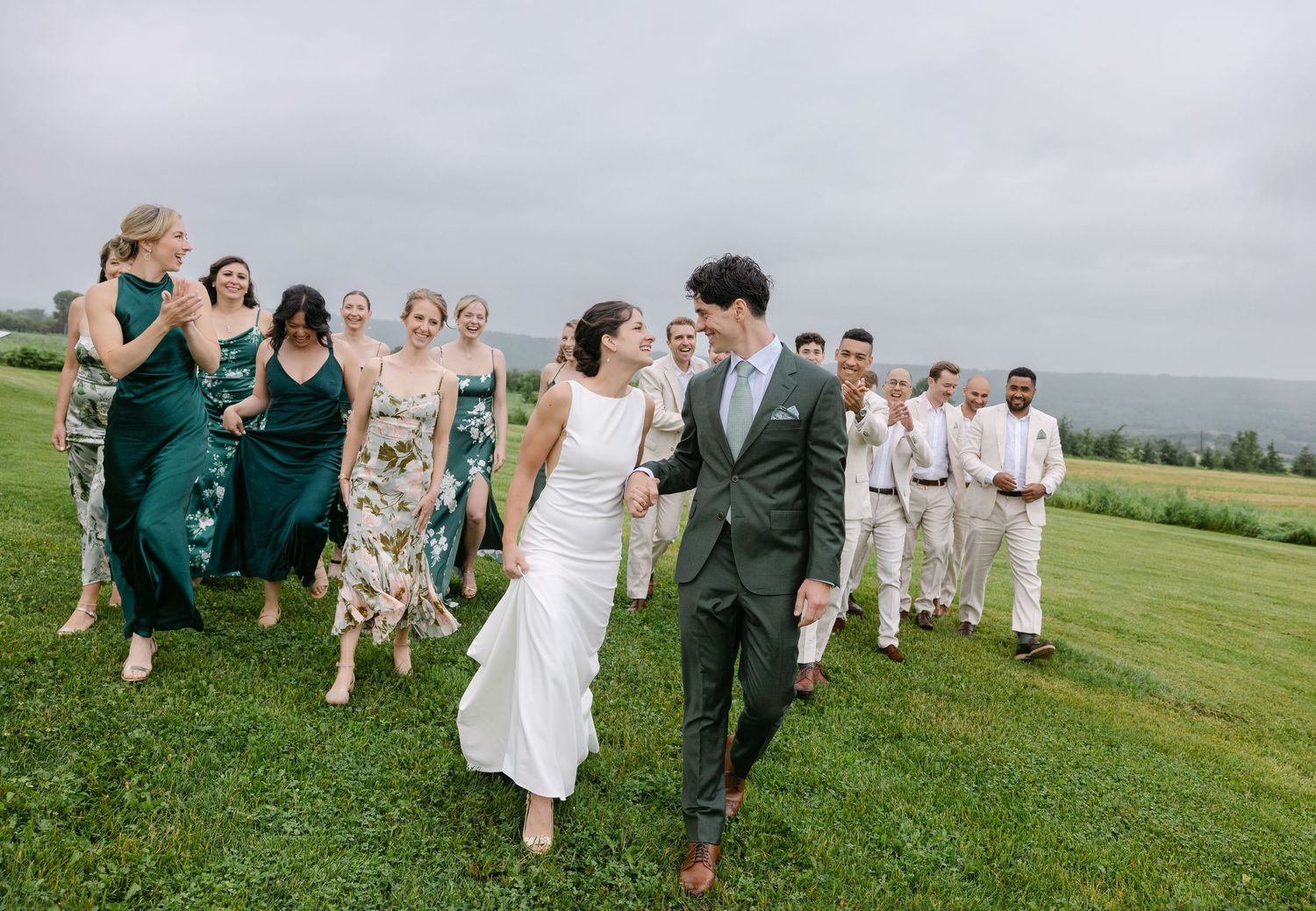Wedding party runs joyfully across a green hillside meadow with mountains in the background on a cloudy day.