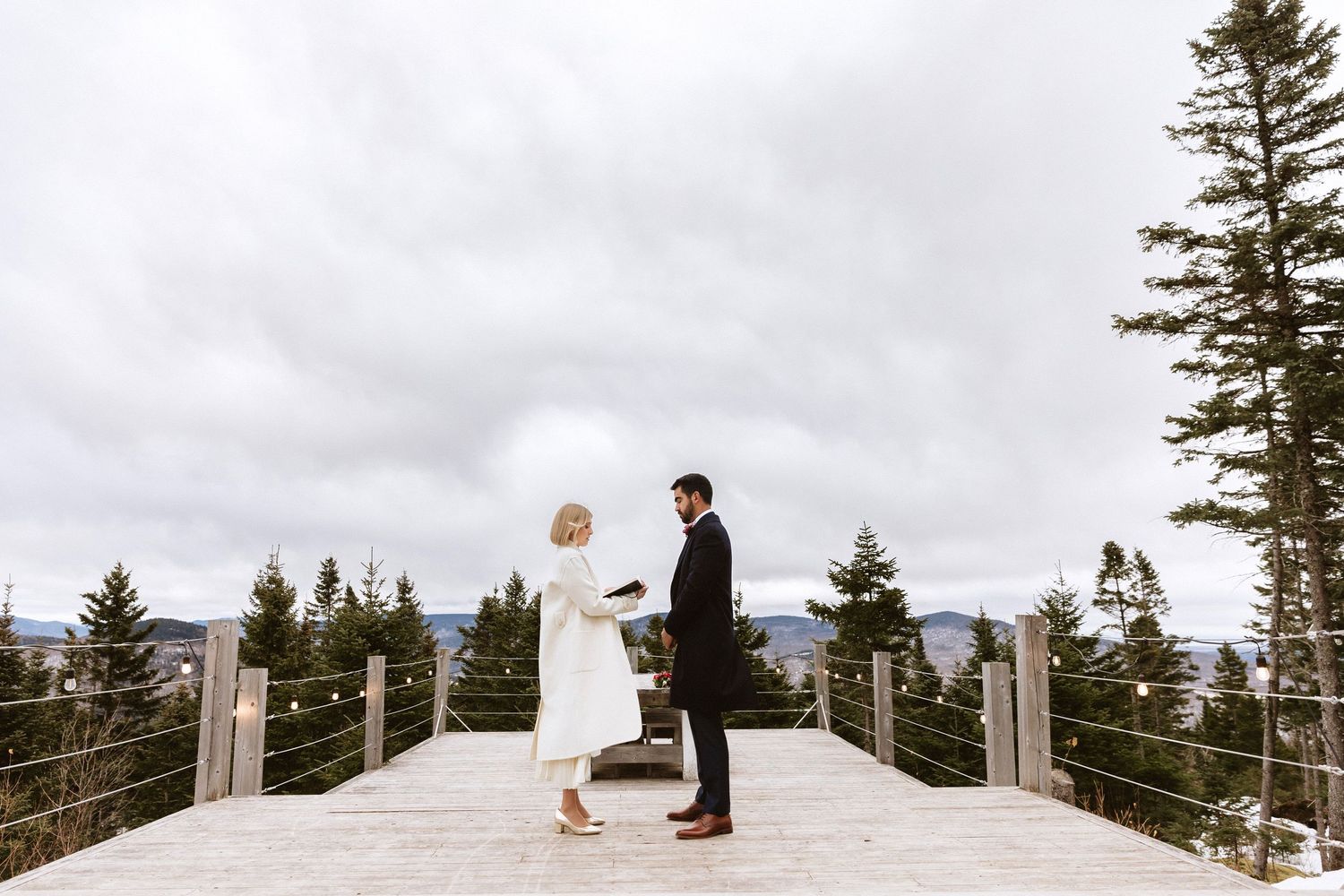 Couple exchanging vows on a scenic mountain deck surrounded by evergreen trees under a cloudy sky.