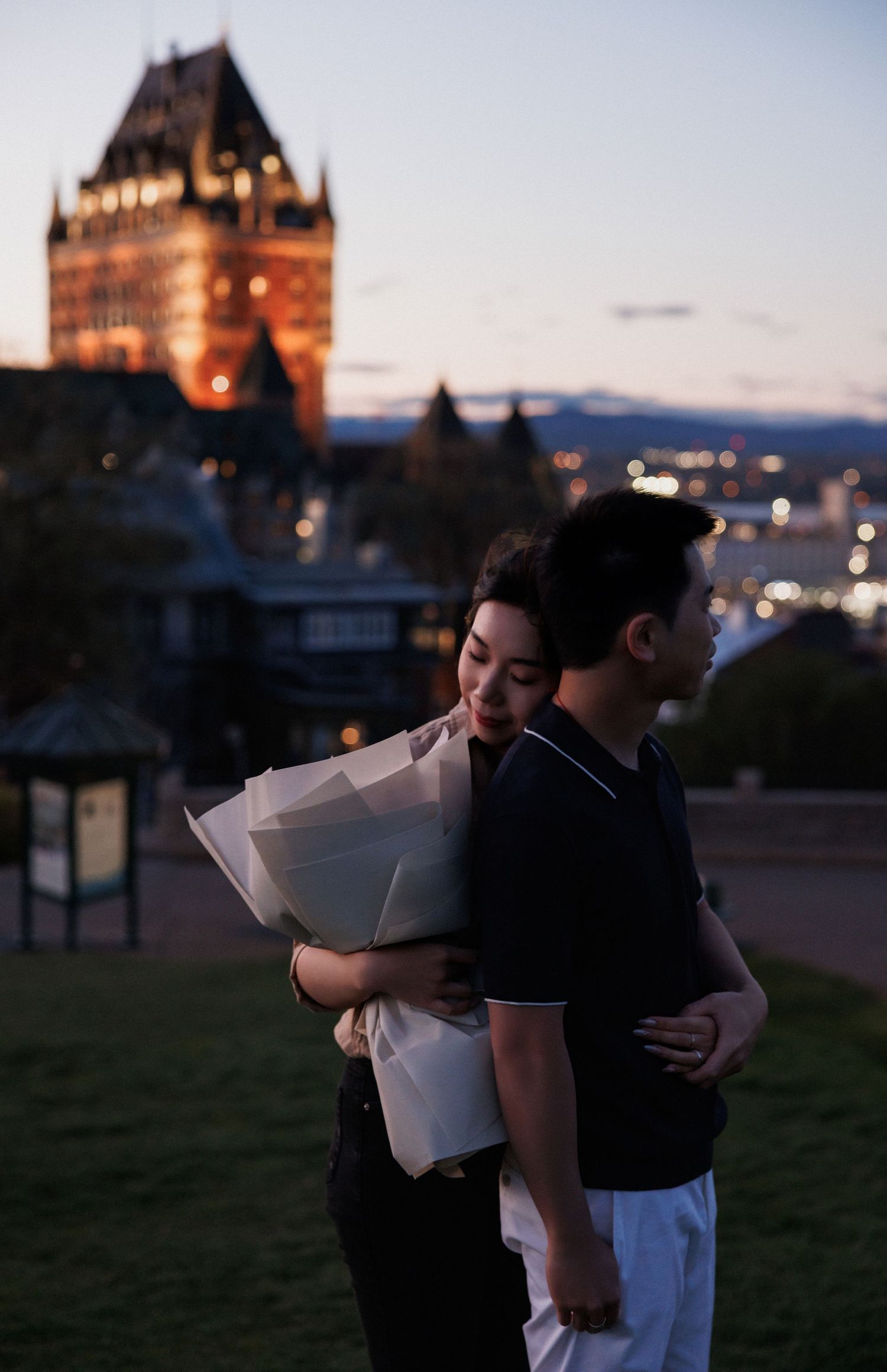 Couple embracing at dusk near Chateau Frontenac in Quebec City, woman holding white flowers.