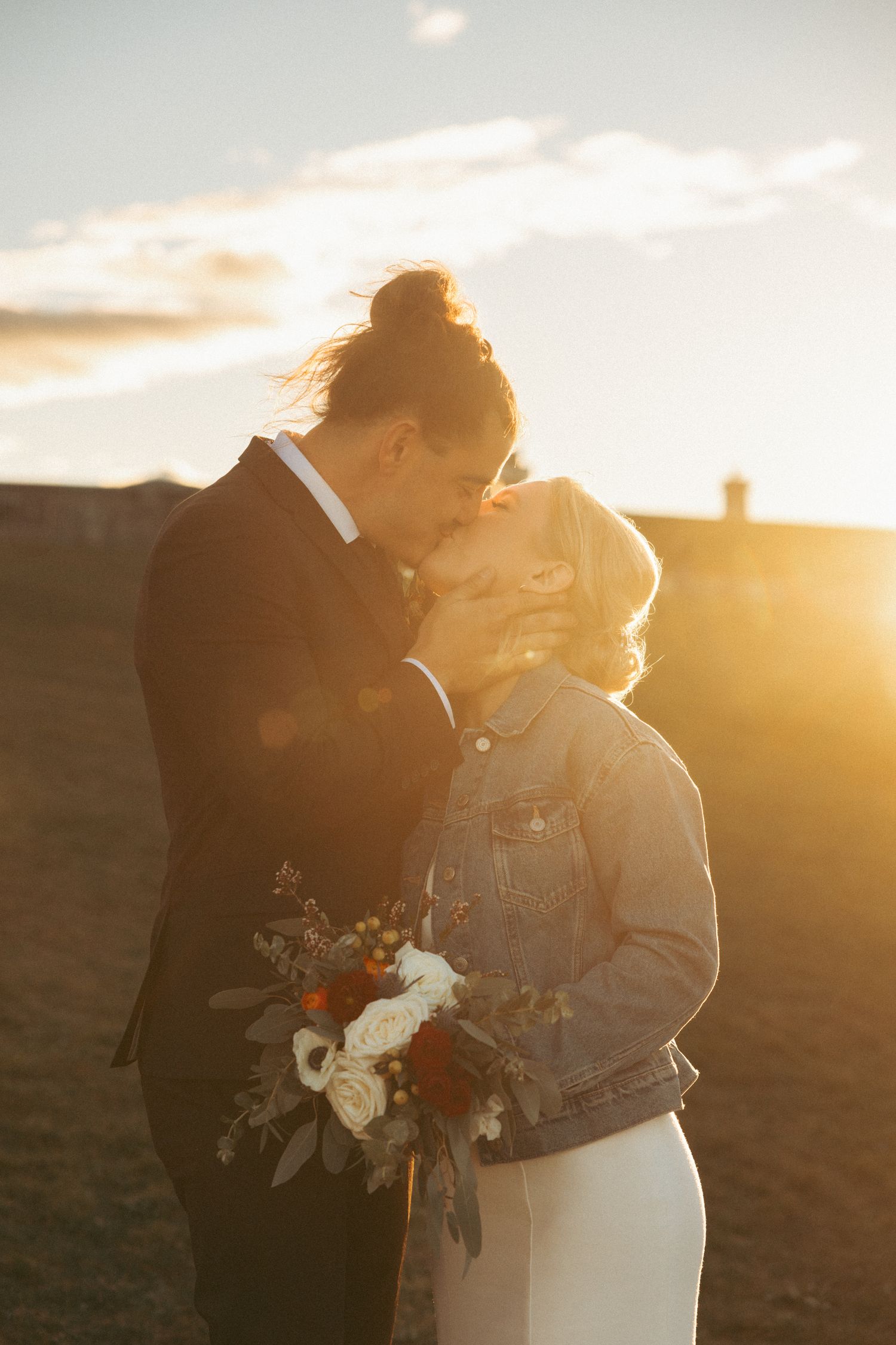 A couple embraces during golden hour sunset while holding a wedding bouquet with white and burgundy flowers.