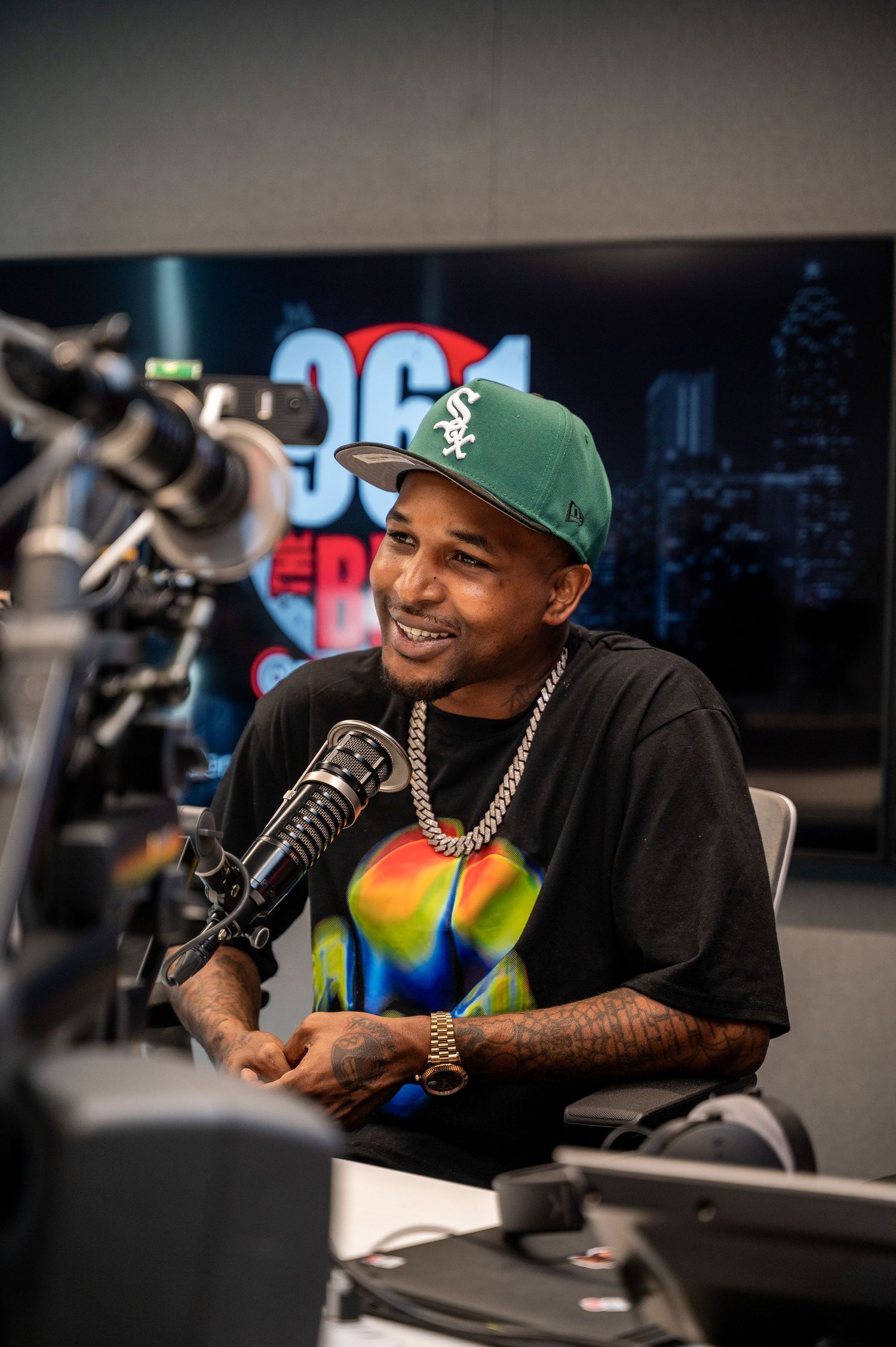 Man in green cap and chain necklace smiling at radio station microphone, wearing colorful graphic tee.