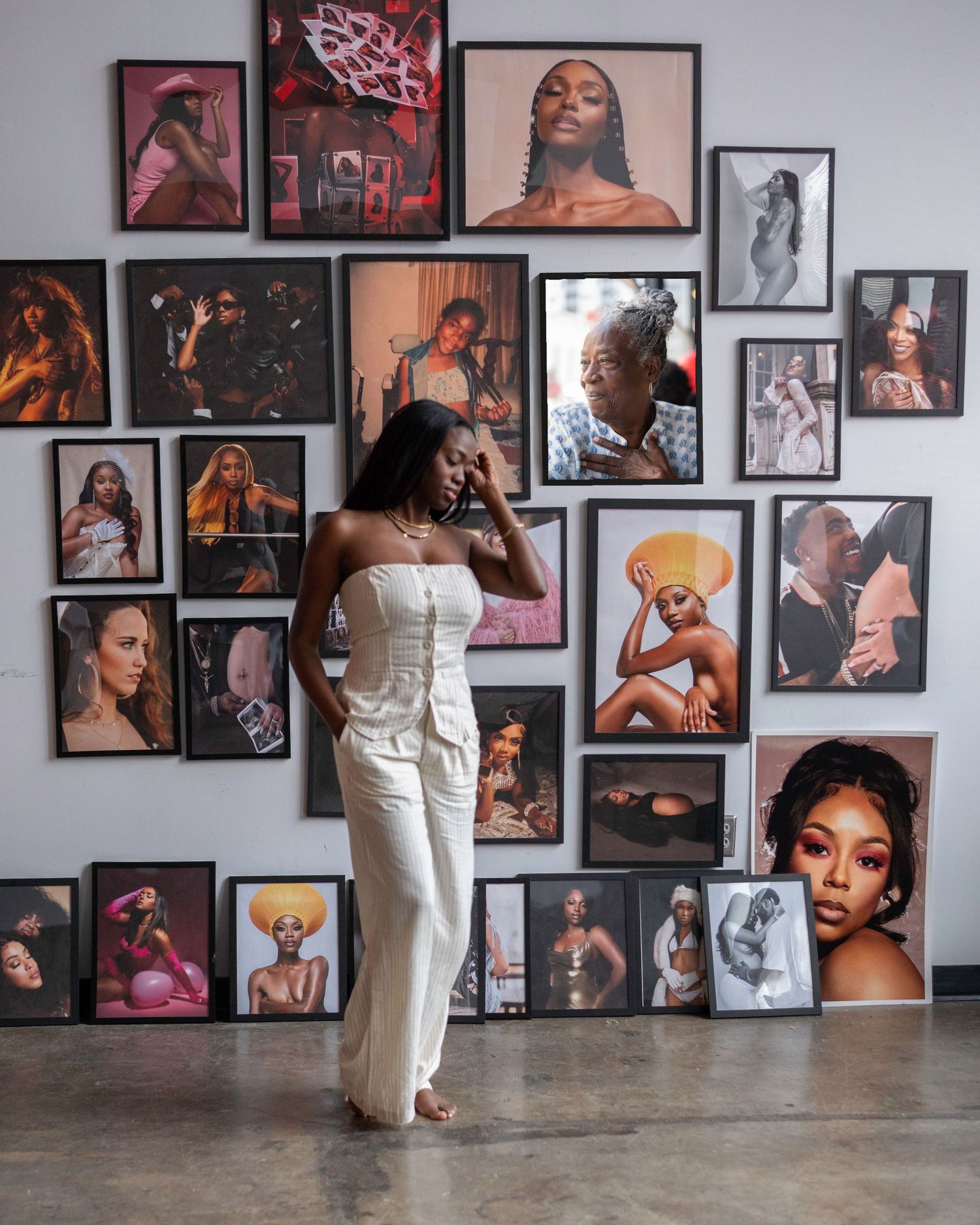 Woman in white outfit poses before a gallery wall of diverse portrait photographs in a modern art space.