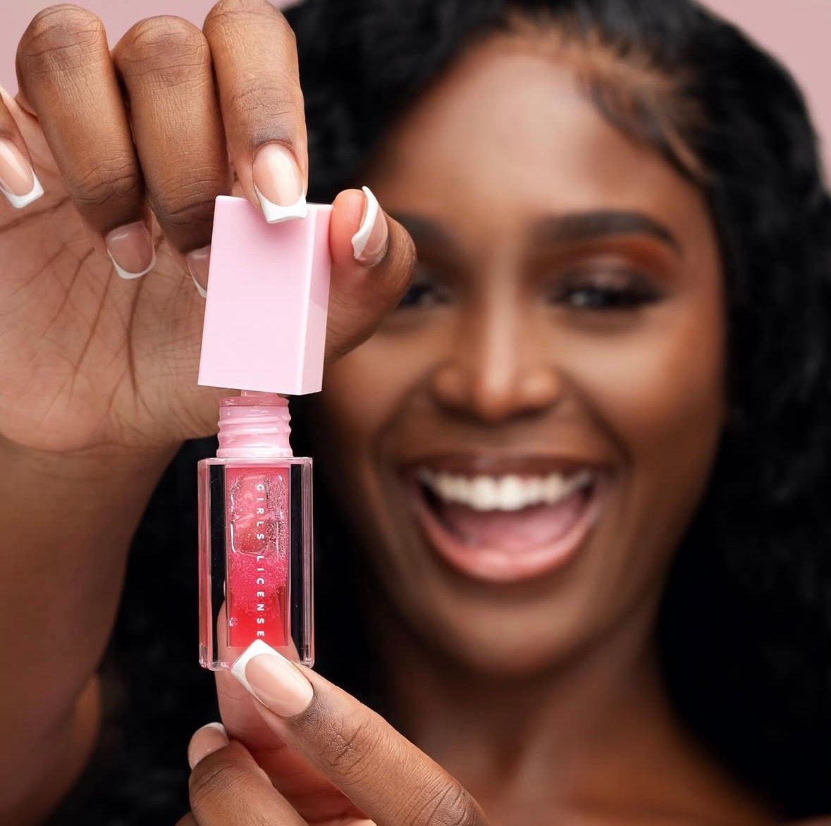 Smiling woman holding open pink lip gloss tube, showcasing sheer pink gloss formula close to camera.