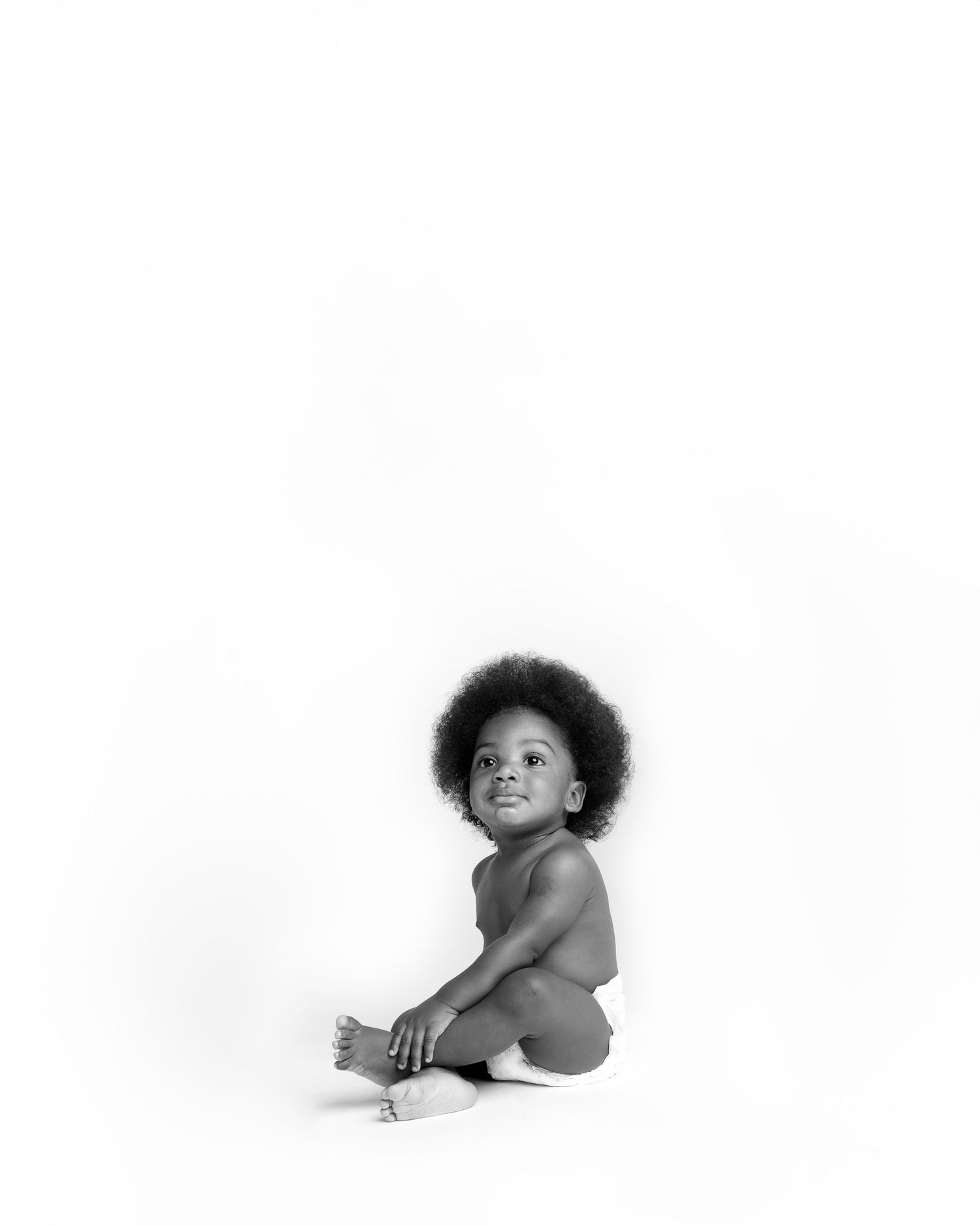 Black and white artistic studio photograph of toddler with afro hairstyle sitting against white background.