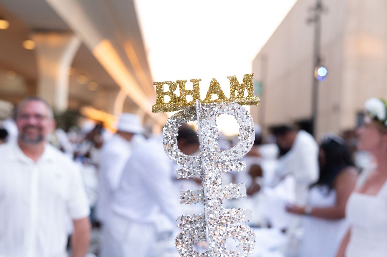 Glittery BHAM sign on decorative stand at elegant white party event with guests dressed in white attire.