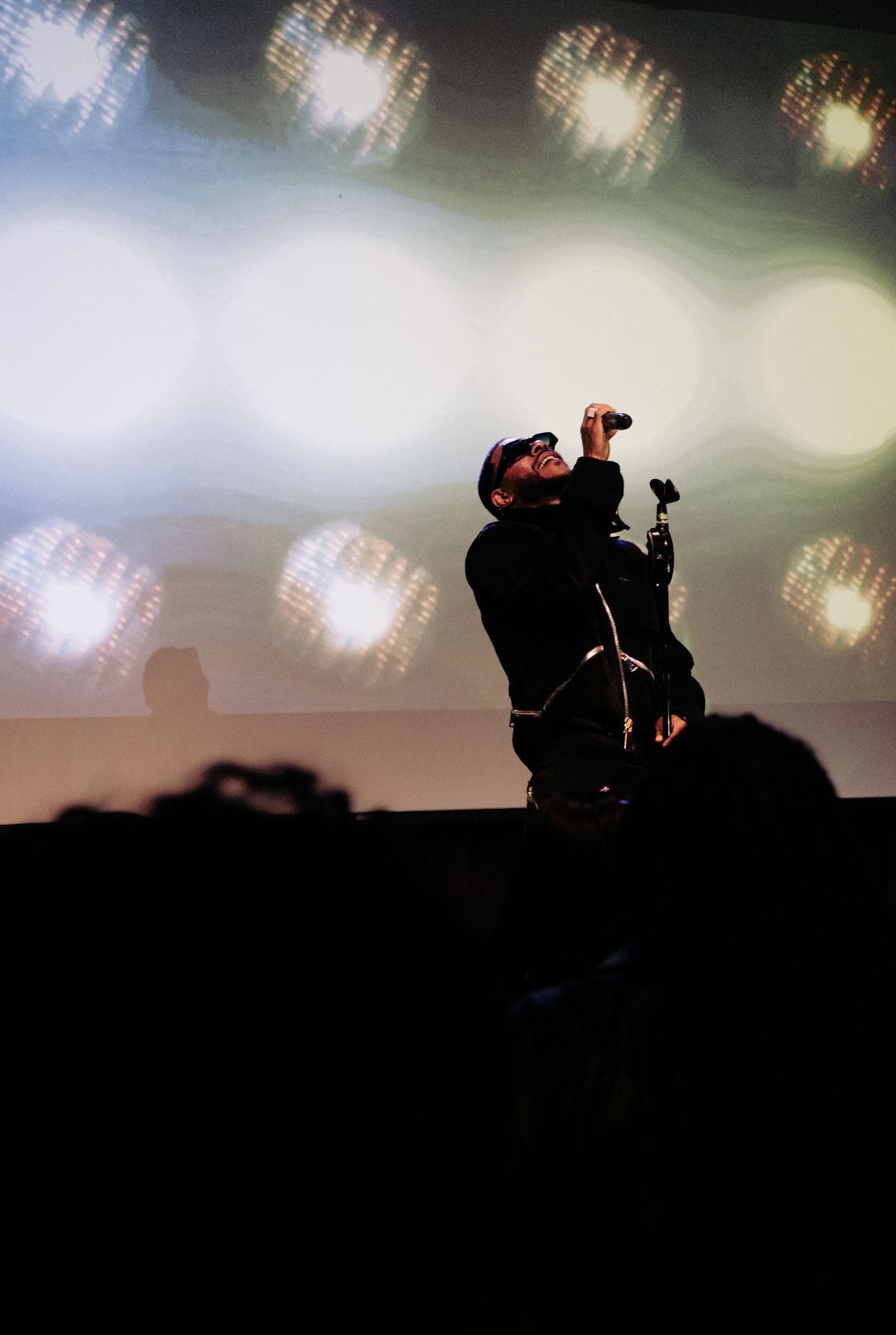 Silhouetted performer singing into microphone on stage with colorful bokeh lights backdrop at live concert.