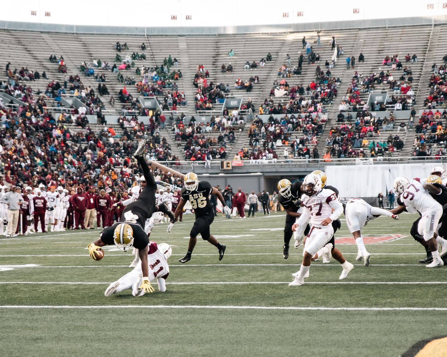 College football game action with players in black and red uniforms competing on the field in a partially filled stadium.