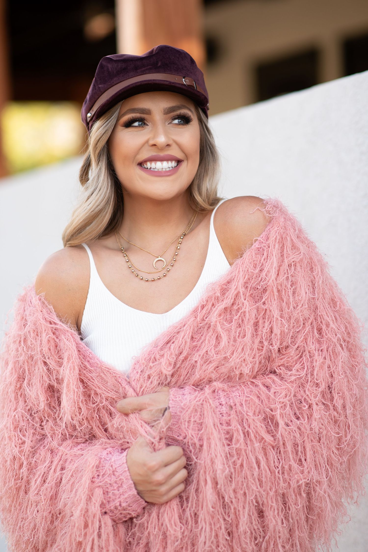 Smiling blonde woman wearing a pink fuzzy cardigan, white tank top, and dark purple newsboy cap with gold necklace.