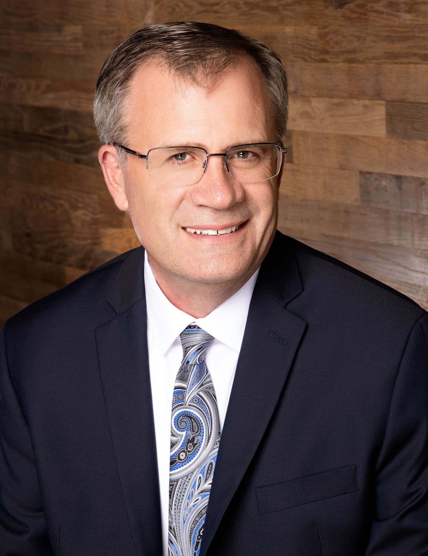 Executive portrait with glasses against wooden paneled wall wearing business attire.
