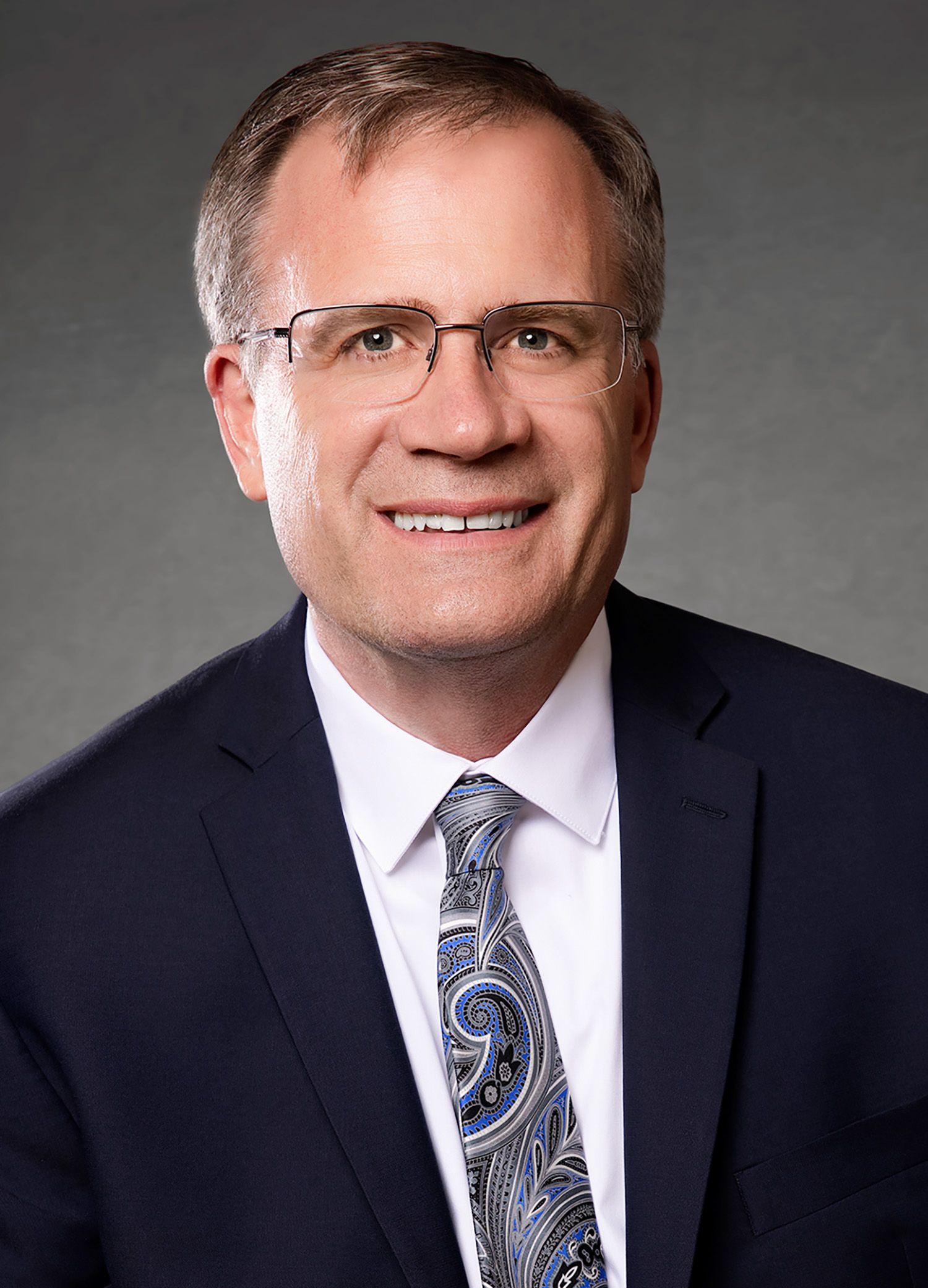Corporate headshot of executive in business attire against grey background.