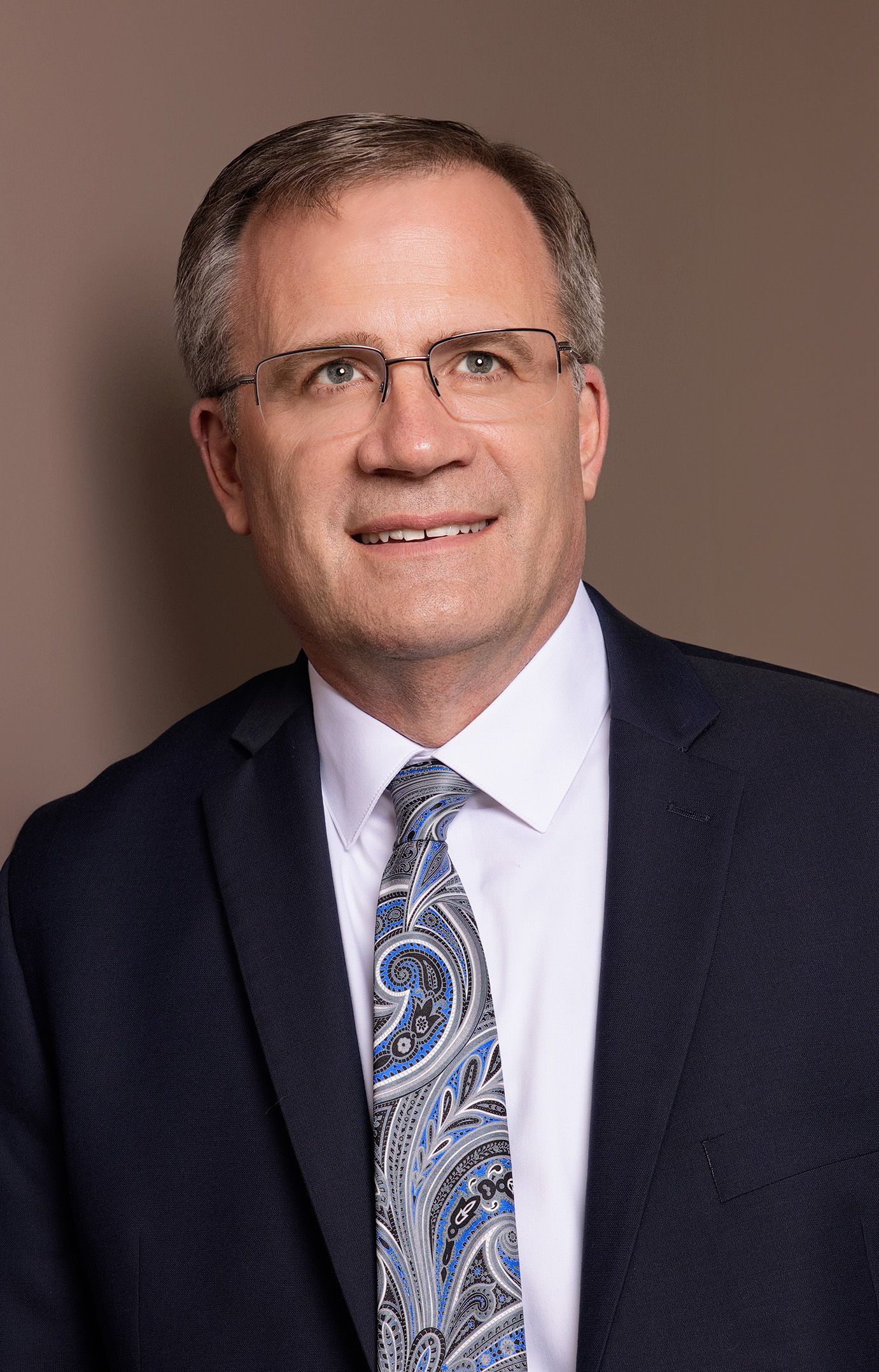 Professional headshot of a business person wearing a navy suit with paisley tie against brown background.