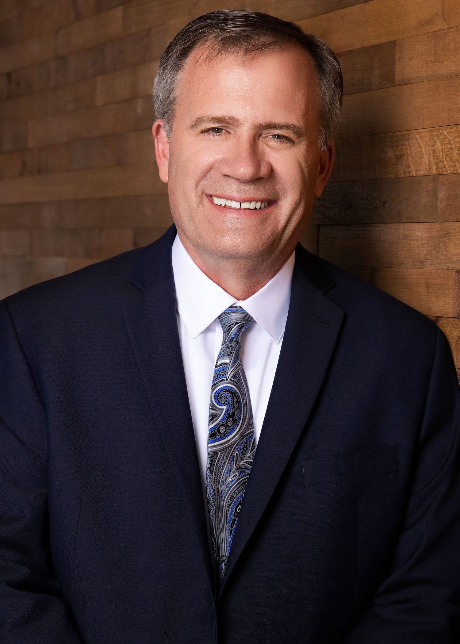 Professional headshot against wooden wall background with navy suit and patterned tie.