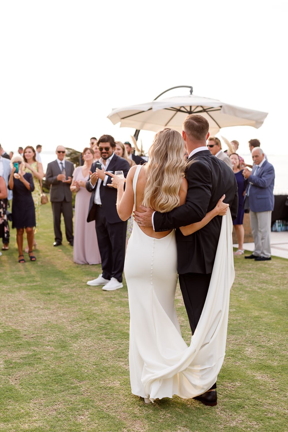 Wedding couple embraced under umbrella surrounded by applauding guests.