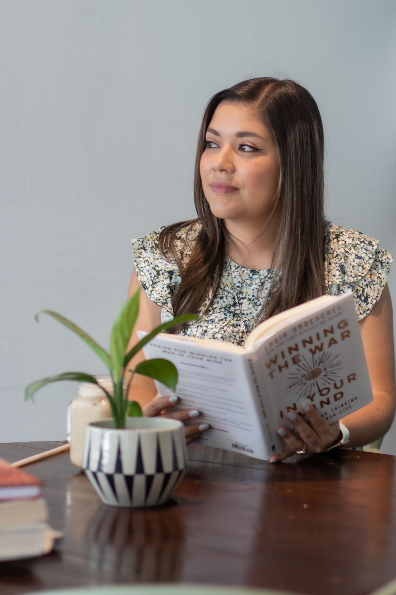 Young woman smiling while reading a book at a wooden table with a small potted plant nearby.