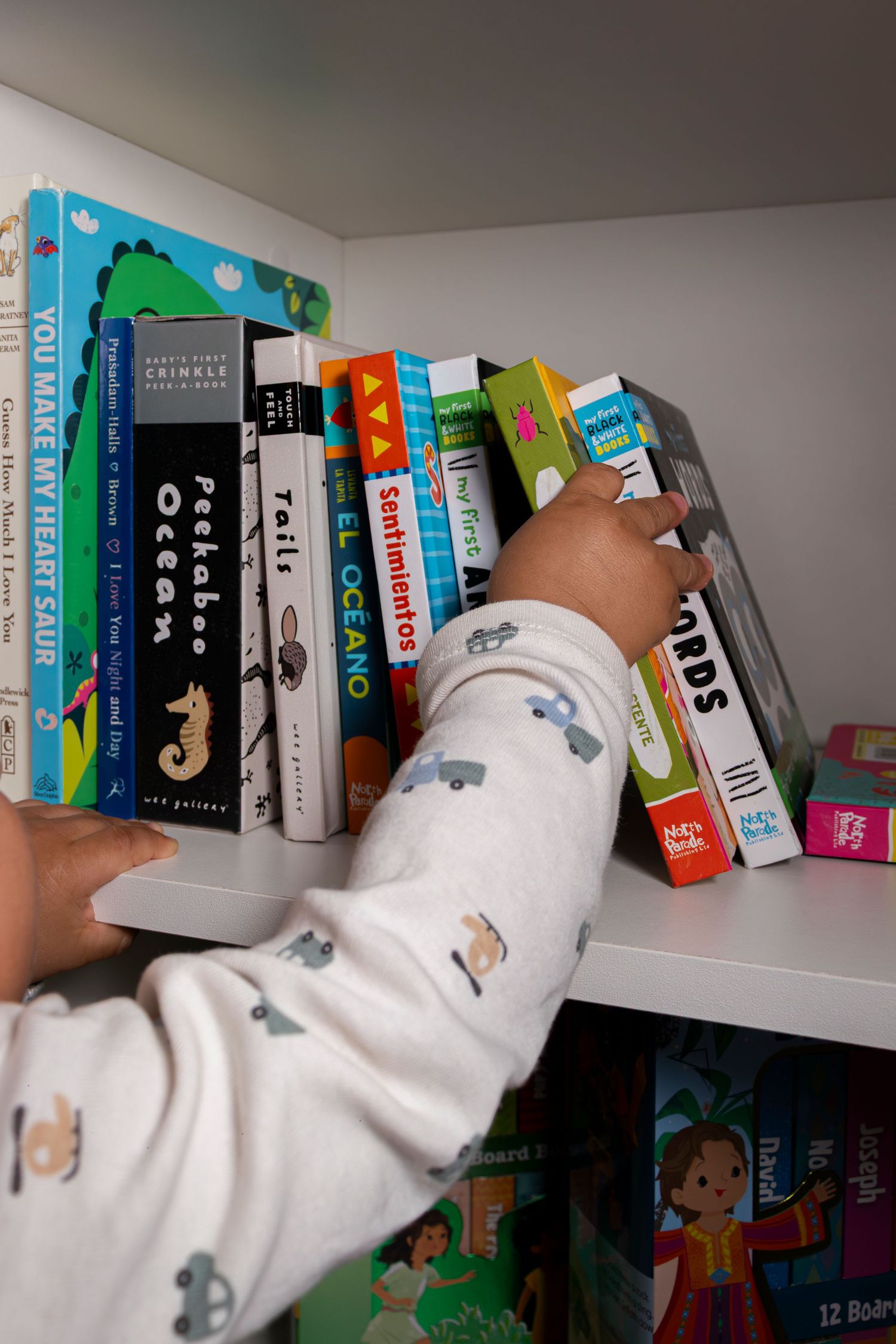 A toddler in a white onesie reaches for colorful children's board books on a white shelf.