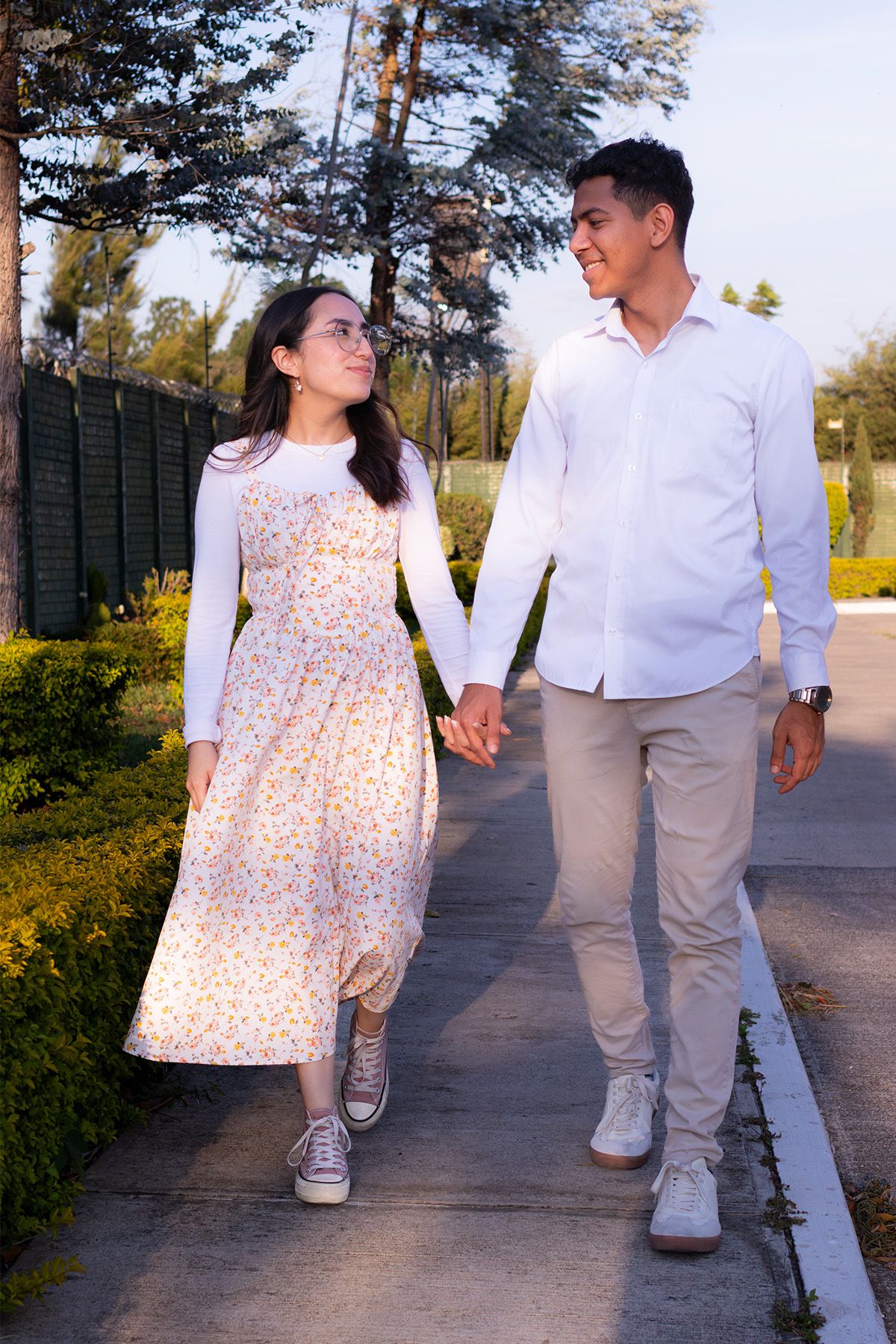 Young couple holding hands and walking together on a garden path, smiling at each other on a sunny day.