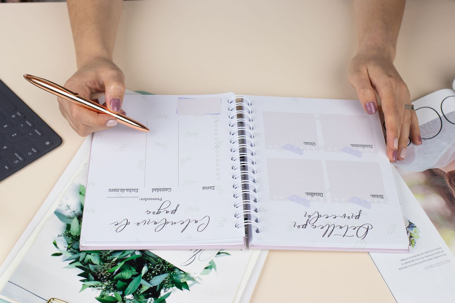Woman writing in an open spiral planner notebook on a desk with a rose gold pen and floral books nearby.