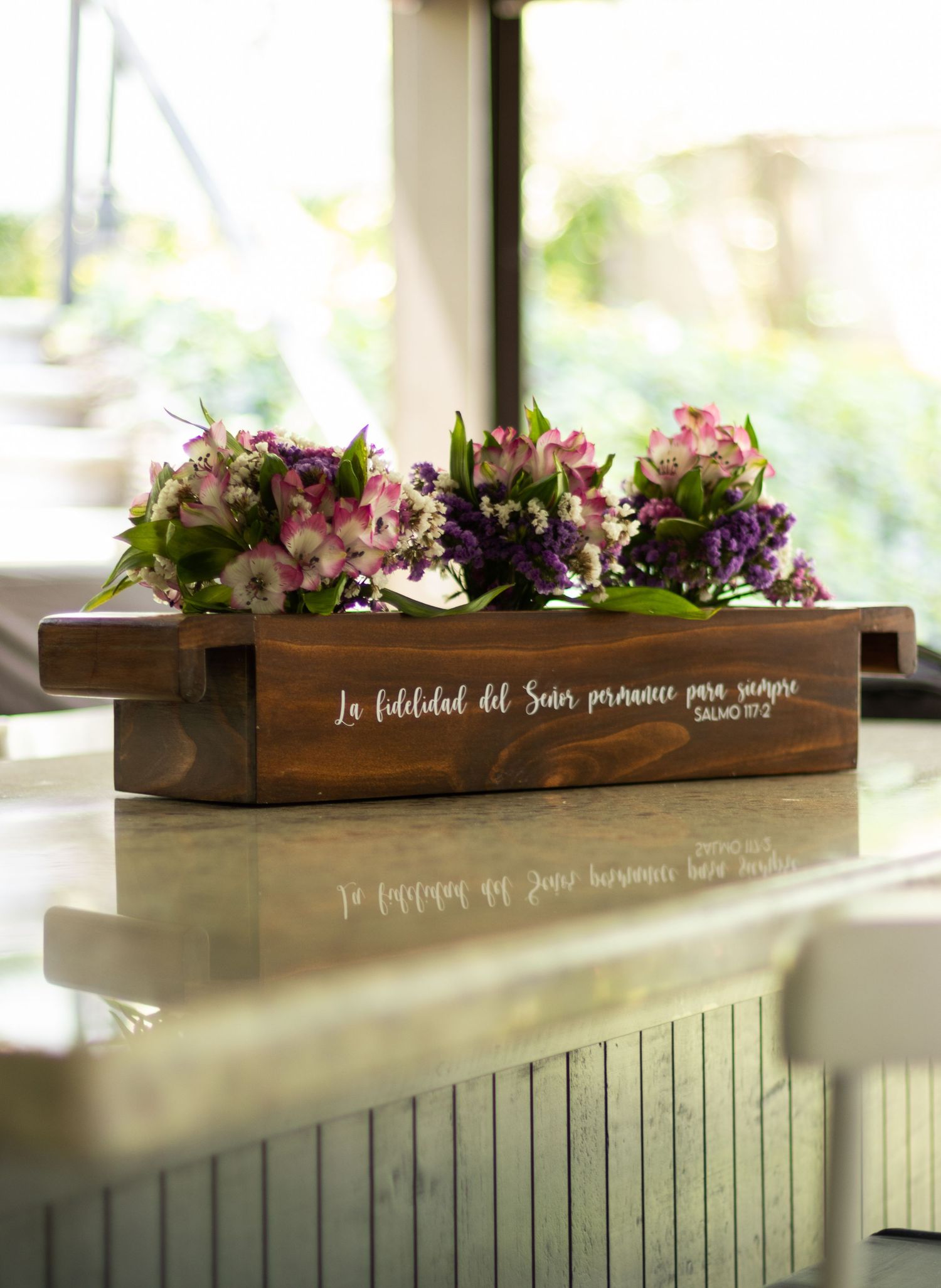 Wooden planter box with Spanish Bible verse holding purple and pink flowers on a marble surface.