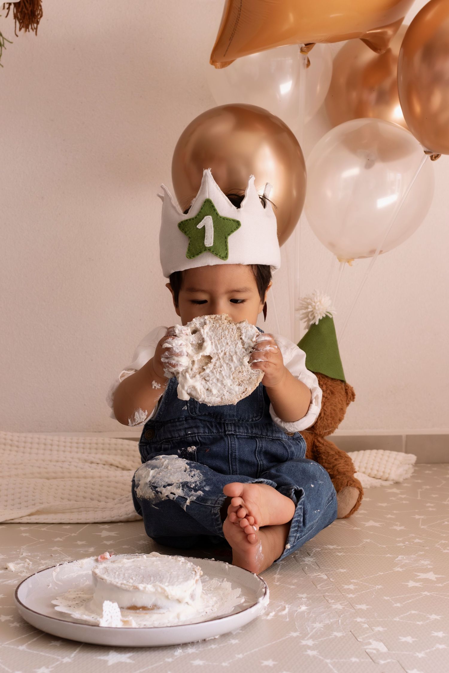 Small child wearing crown and overalls enjoys messy cake smash photo session with white and gold balloons in background.