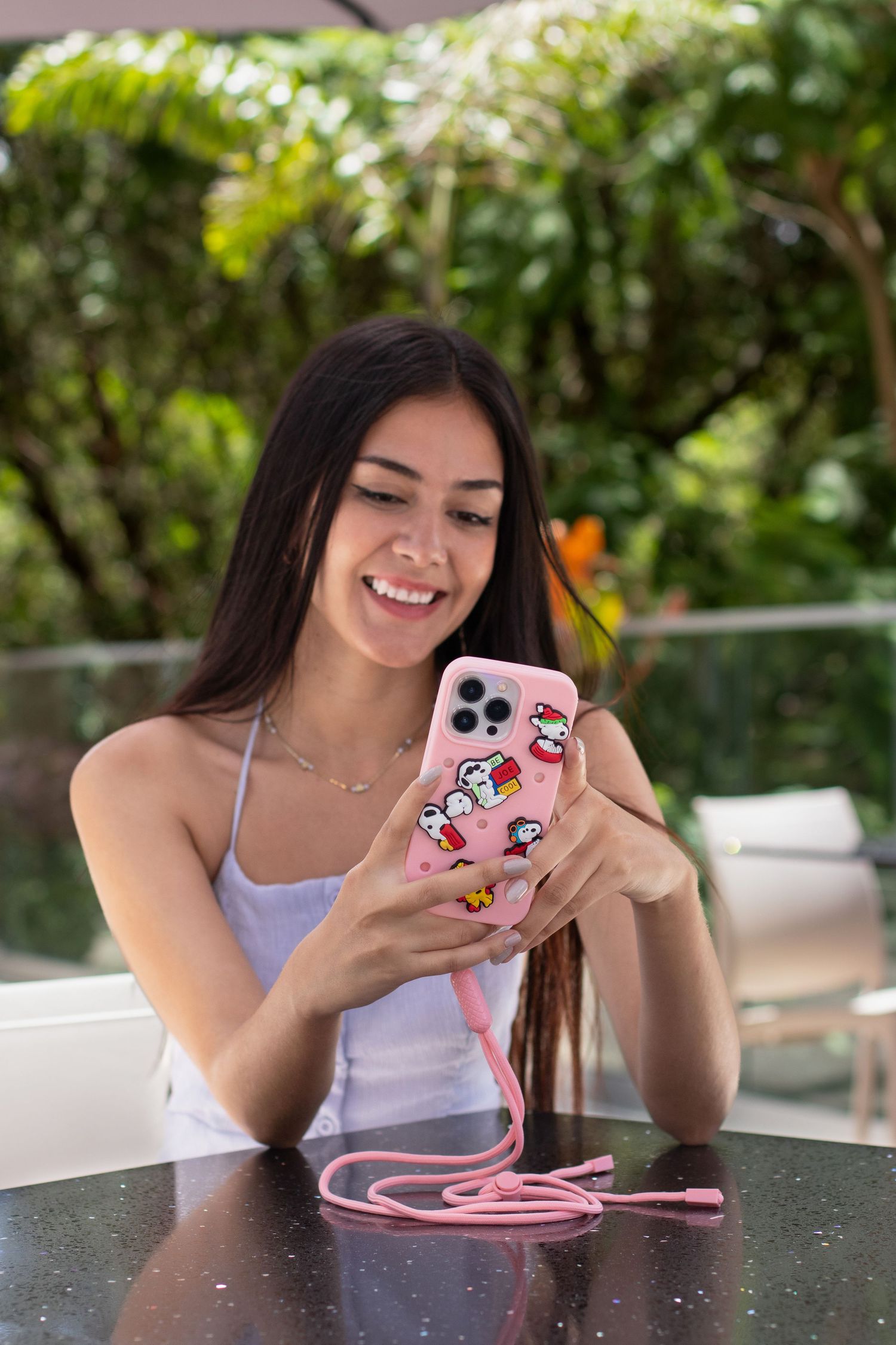Person using a pink phone case with lanyard while sitting outdoors at a table.