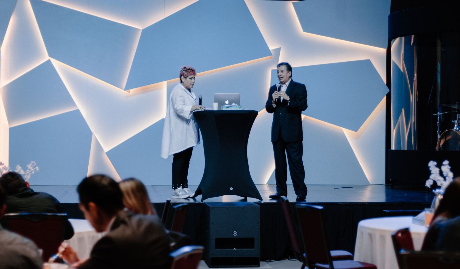 Two speakers present on a modern geometric lit stage at a professional conference event.
