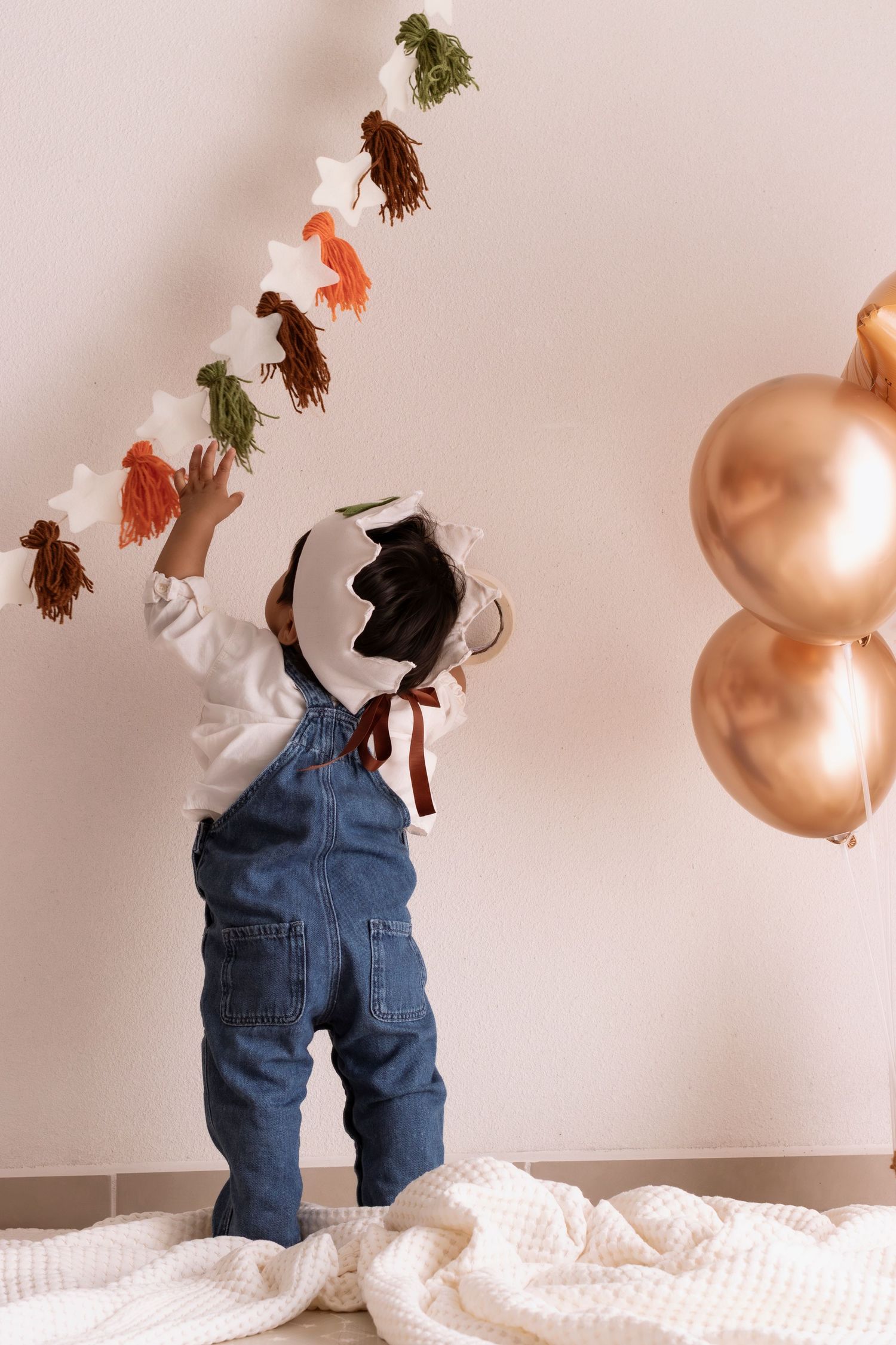 A baby wearing denim overalls crawls on a white textured surface while colorful toys dangle overhead.
