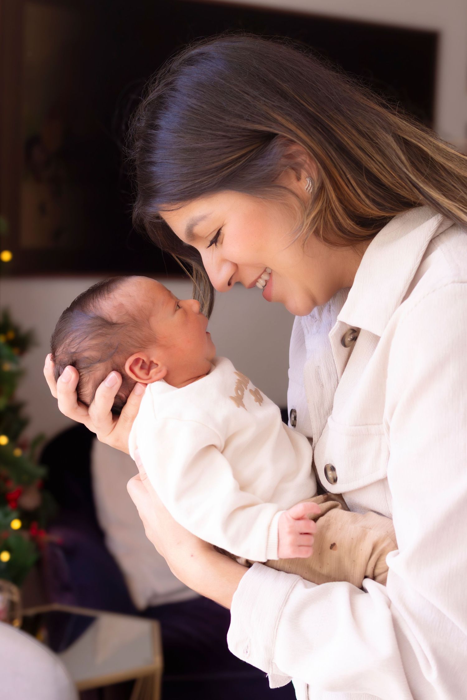A smiling mother tenderly holds her newborn baby near a Christmas tree, both dressed in white.
